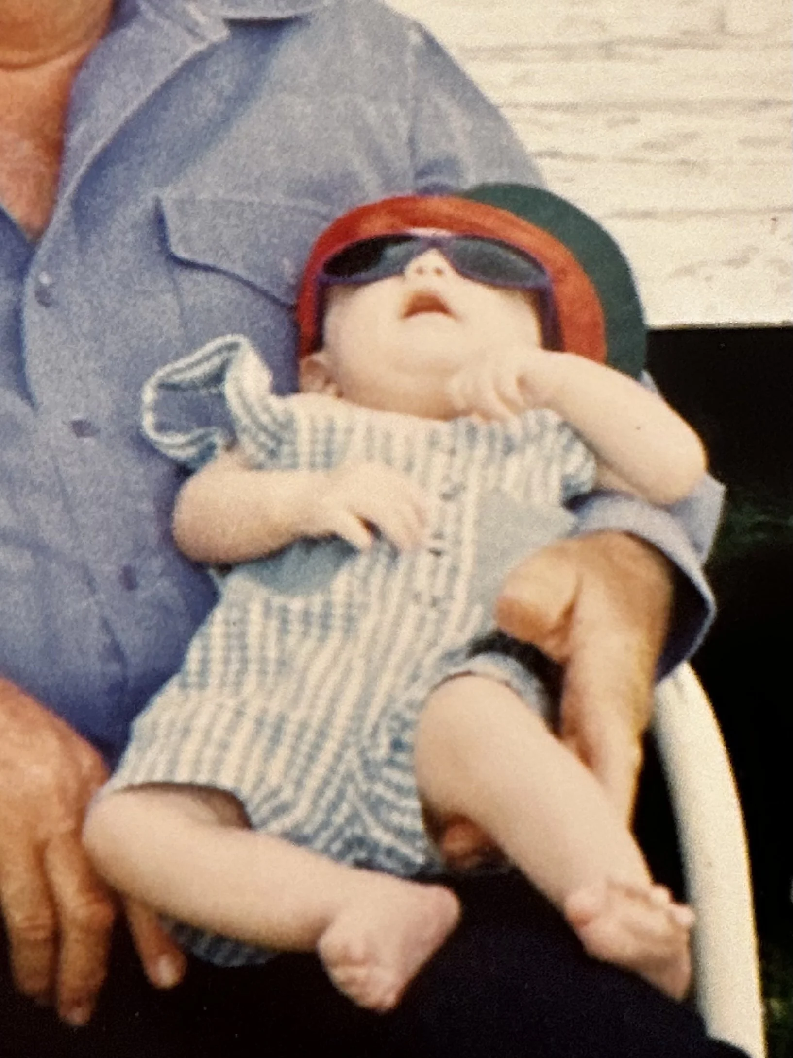A person holding a baby in their arms. The baby is wearing sunglasses and a striped outfit, and appears to be relaxed or sleeping.