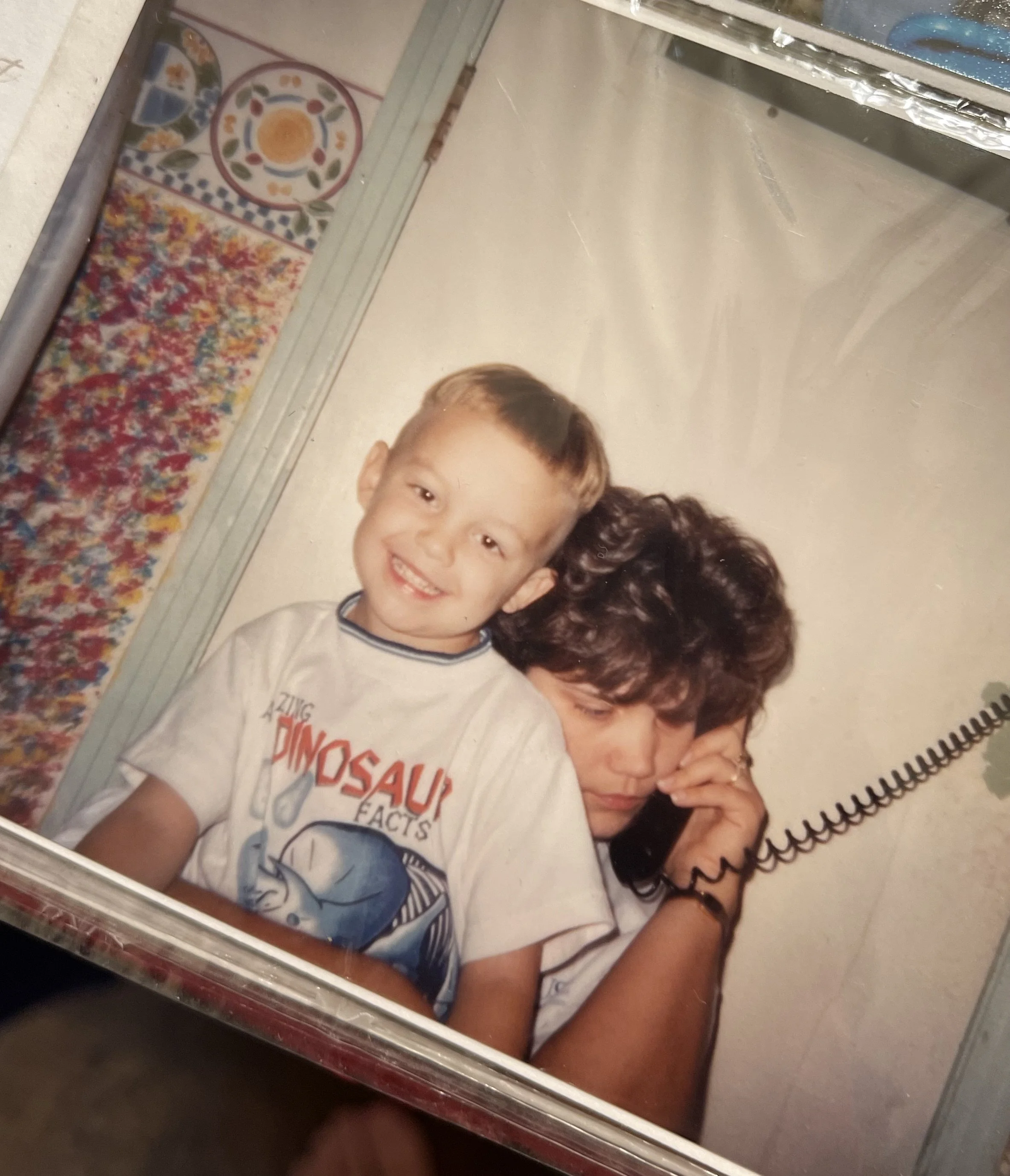 Printed photo in an album. A young boy smiling, wearing a white T-shirt with a graphic and text, and sitting close to an adult woman with dark curly hair. She holds a landline phone to her ear.