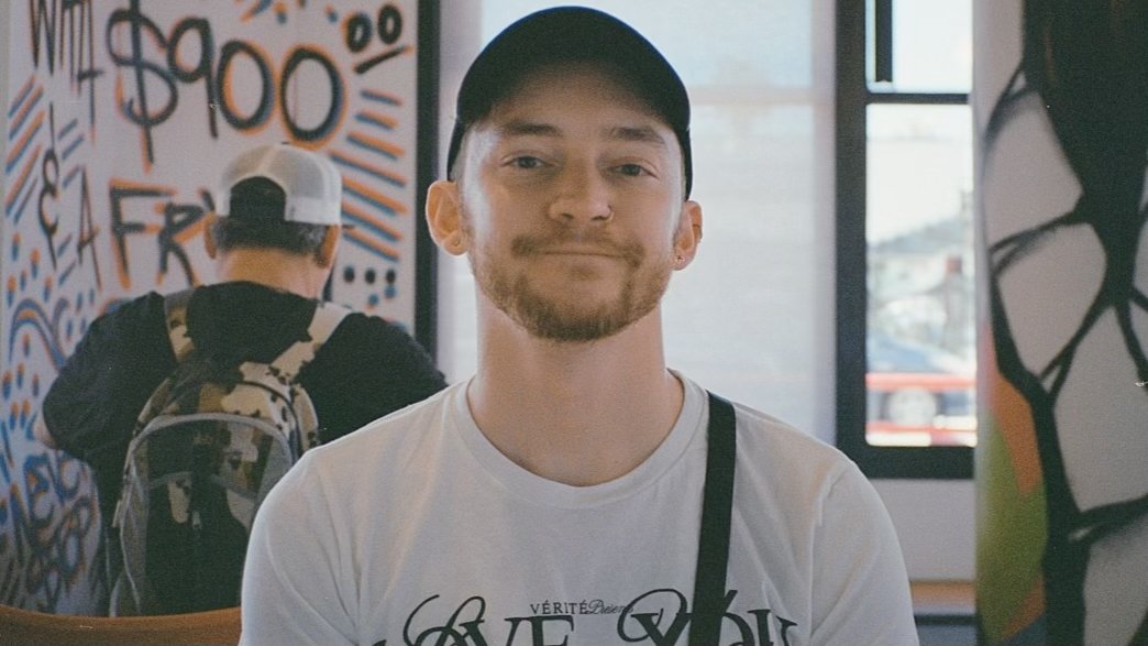 A young man with a beard and a black baseball cap sitting indoors, smiling at the camera. In the background, there is a person with a white cap and a backpack, and colorful graffiti on the wall.