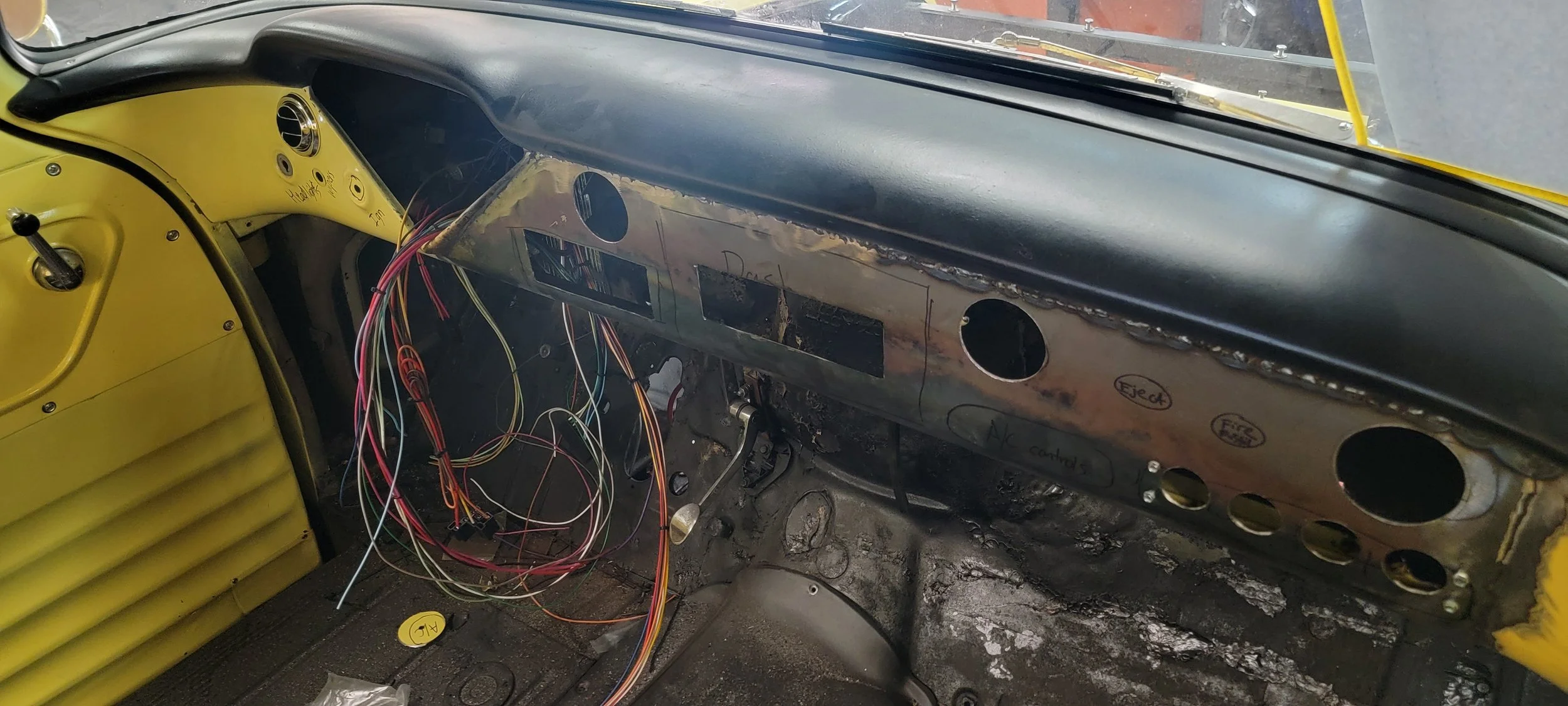 The interior of a vintage race car in progress of restoration, showing a yellow dashboard, exposed wiring, and a rusted metal dashboard with handwritten labels.
