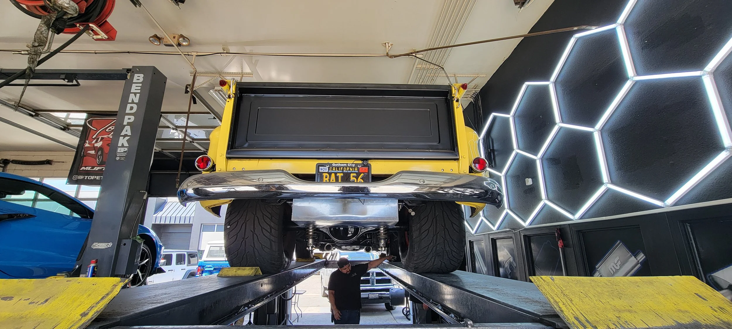 A yellow vintage car is lifted on a hydraulic lift inside a garage. The car has a California license plate with the text 'BAT 56' and Gotham City written on it. There are neon hexagon wall lights and a mechanic working underneath the car.