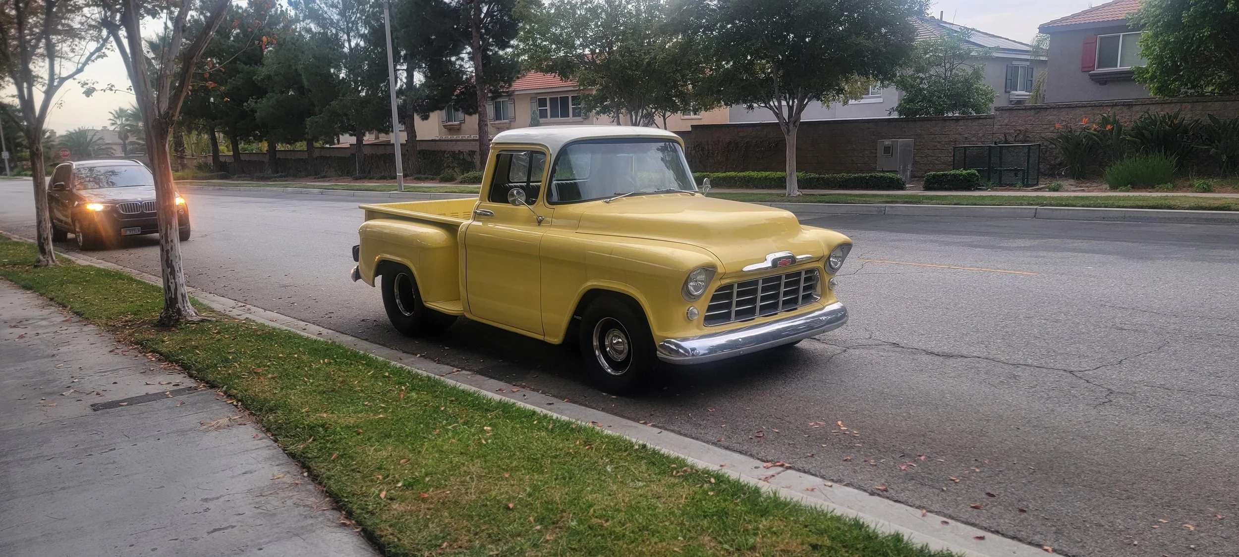 A yellow vintage pickup truck parked on the side of a residential street with trees and houses in the background.