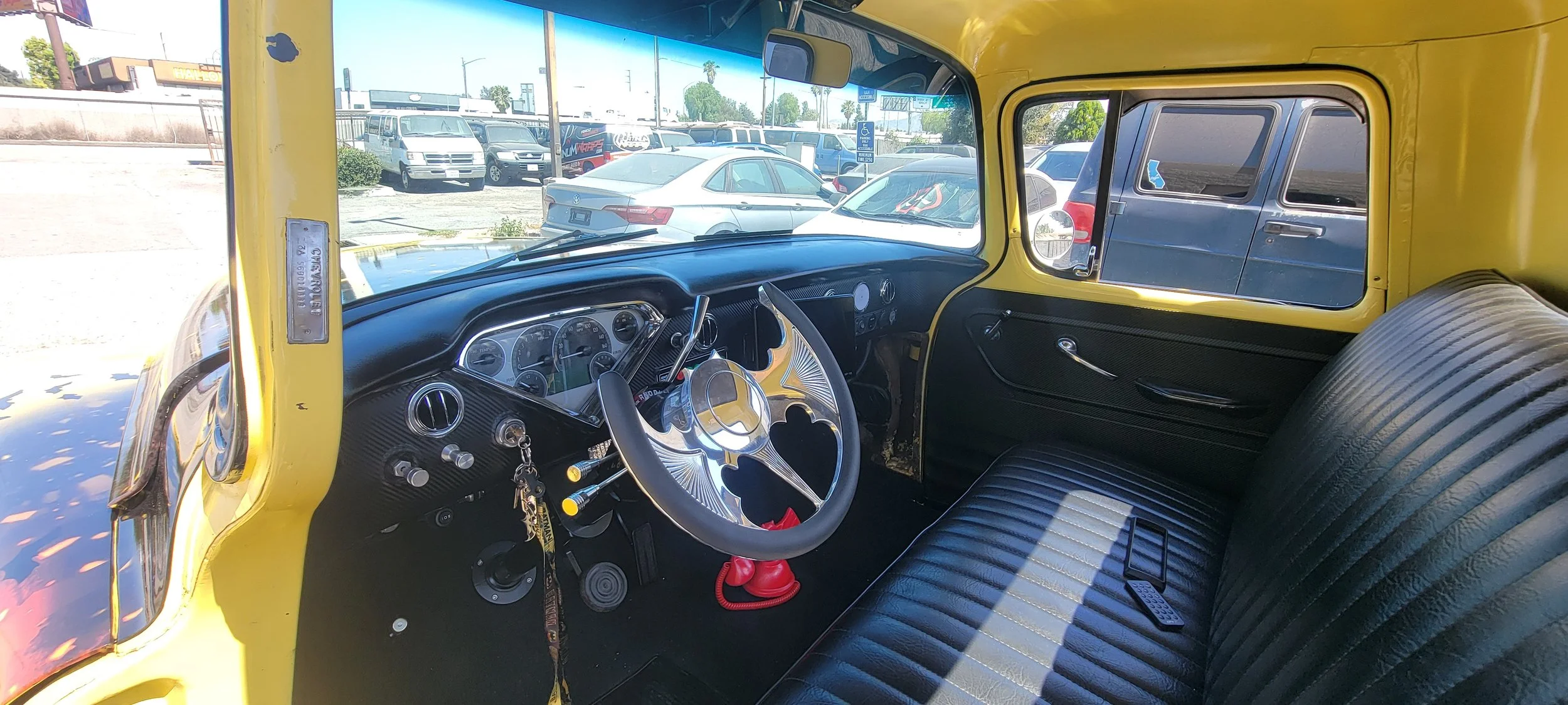 Interior of a vintage yellow truck with a black dashboard and black seat upholstery; there are keys in the ignition, a decorative steering wheel, and a red fire extinguisher on the floor; outside the truck, a parking lot with various cars and a sign 