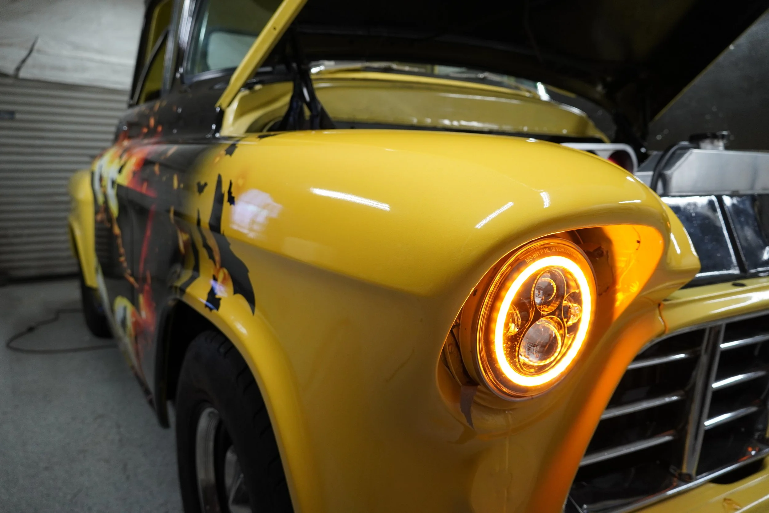 Yellow customized car with bright orange glowing headlights, hood open, in a garage or workshop.