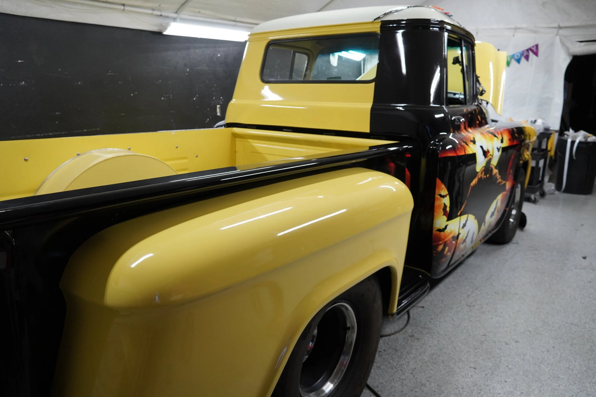A yellow and black vintage pickup truck with a fiery graphic design on its side, parked indoors in a garage or workshop.