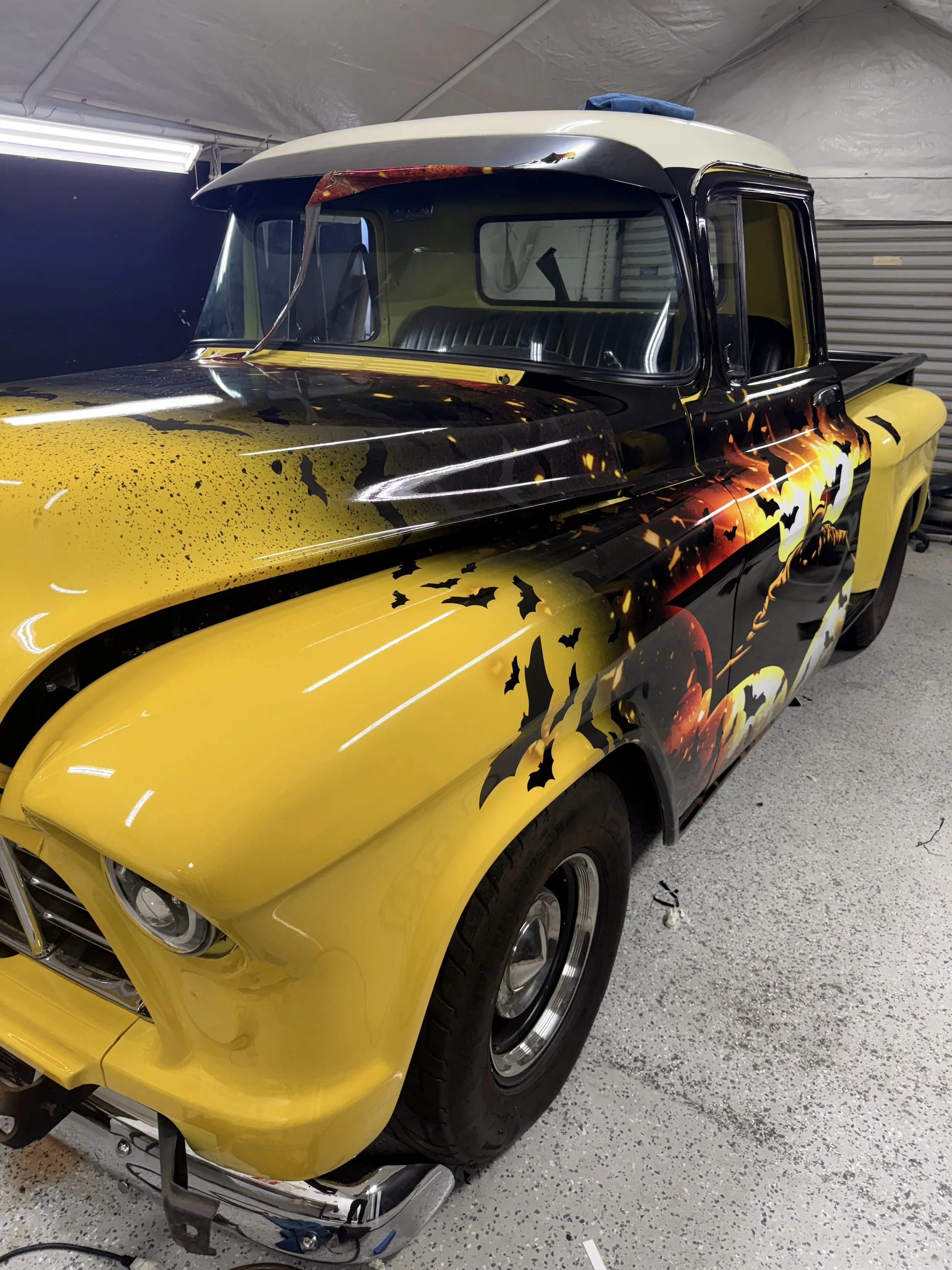 A classic pickup truck painted with Halloween-themed graphics, including bats, pumpkins, and a night scene, parked inside a garage with a speckled floor.