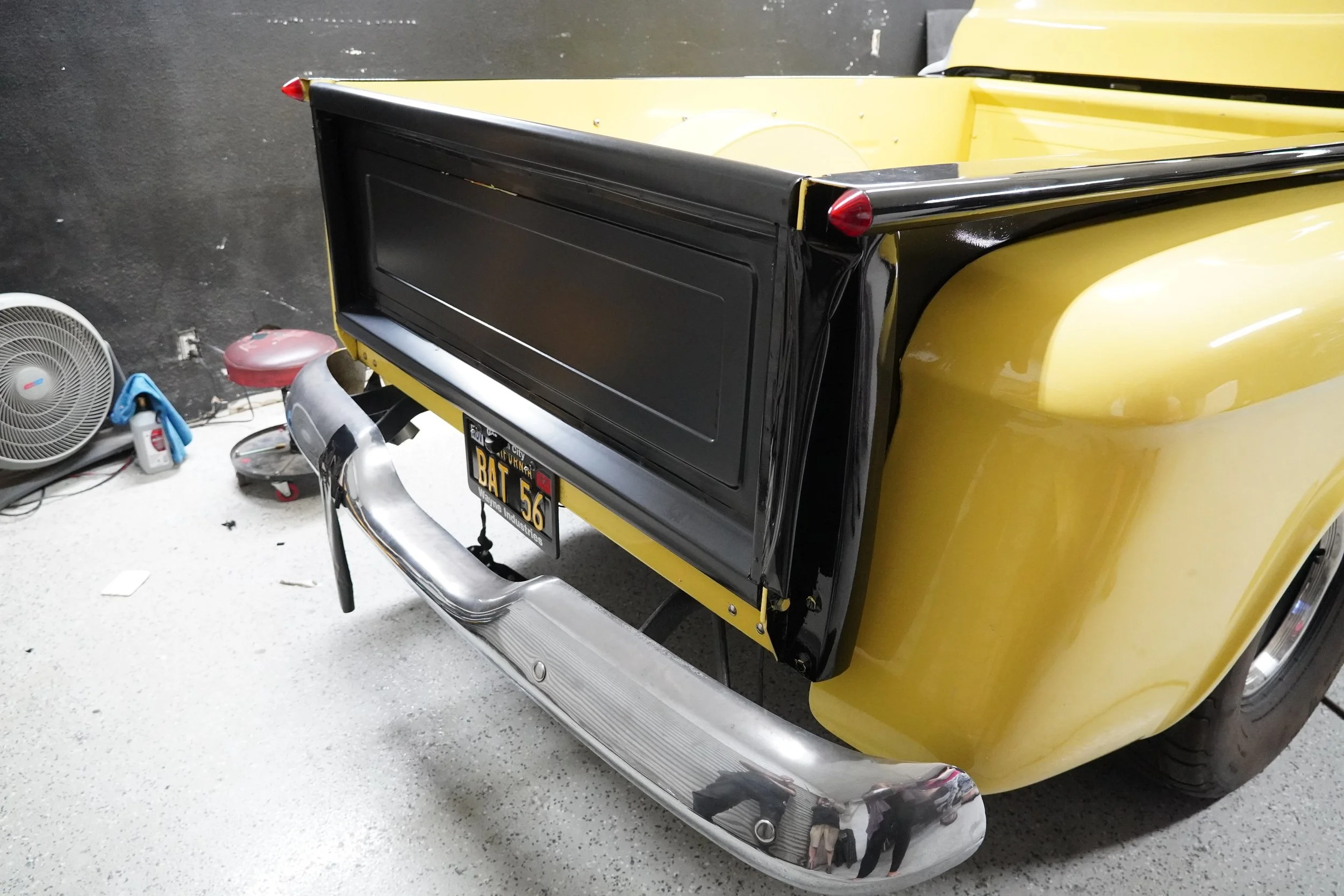 Close-up of the rear of a vintage yellow car with black trim, tail lights, chrome bumper, and license plate reading 'BAT 56'. The background shows a black wall, a floor fan, cleaning supplies, and a red stool.