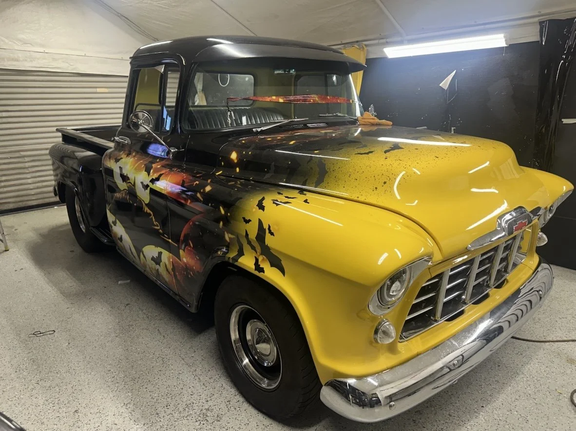 A vintage pickup truck with a black and yellow paint job featuring a bat and moon design, parked in a garage.