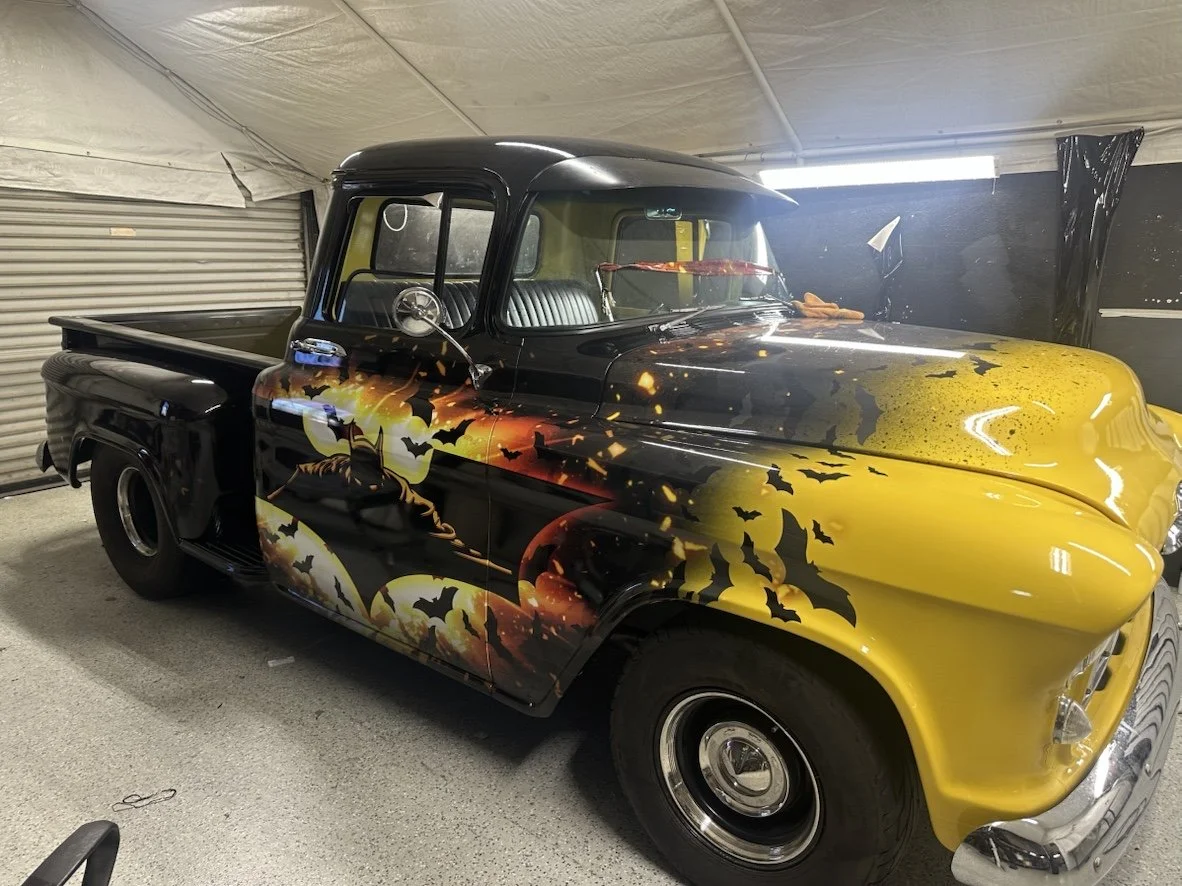 A vintage pickup truck with a Halloween-themed airbrushed mural, featuring bats, a full moon, and a haunted house scene, parked inside a garage.