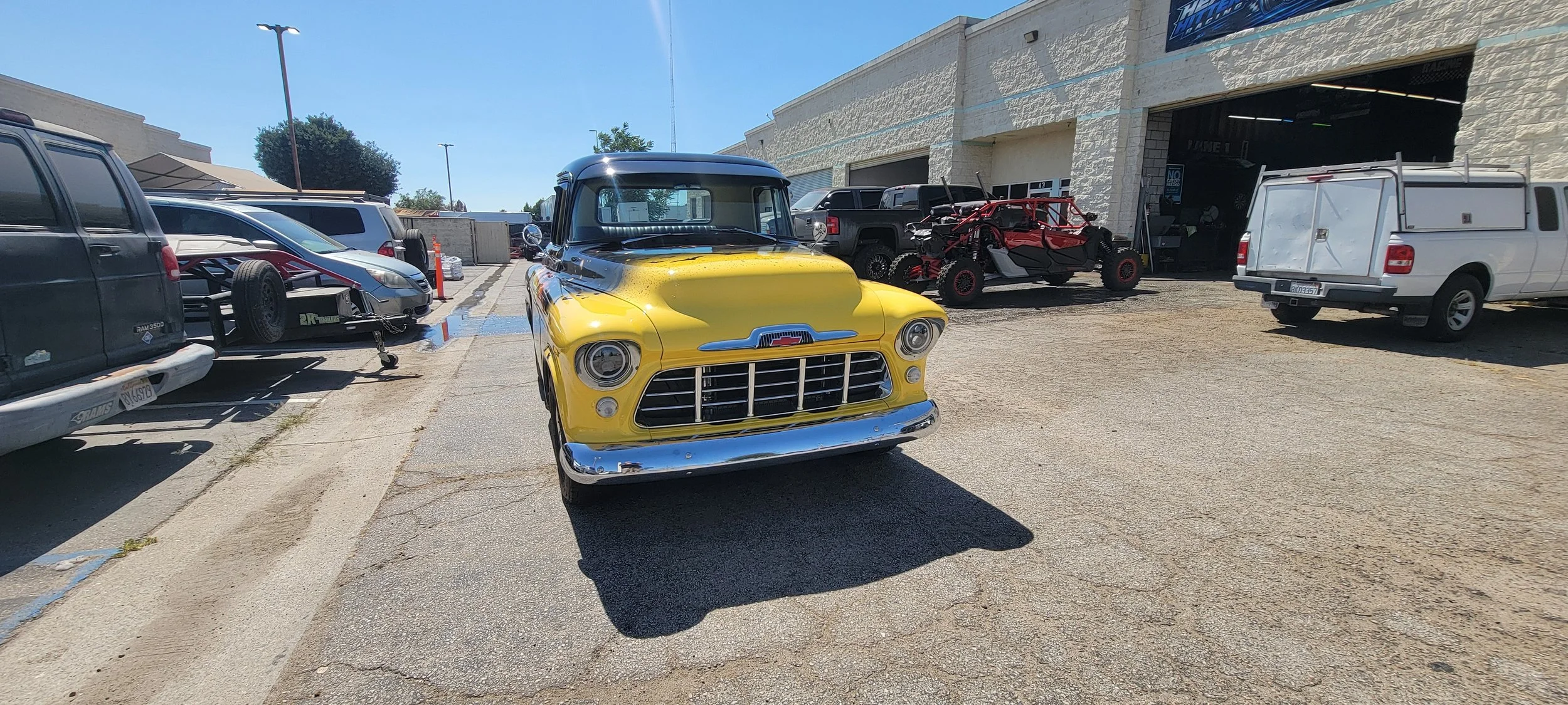 A parking lot outside a building with various vehicles, including a yellow vintage truck, vehicles on a trailer, and off-road vehicles. The building appears to be an auto shop or garage.