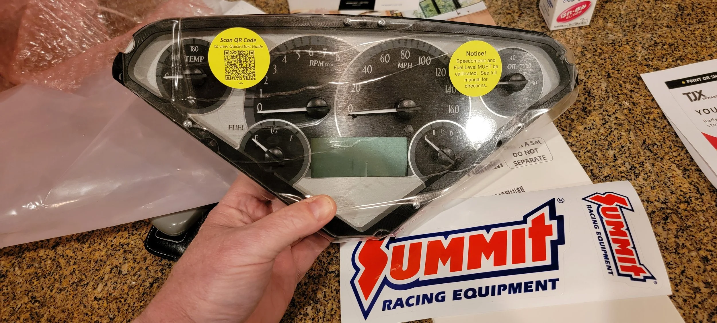 A car dashboard instrument cluster with various gauges, including speedometer, tachometer, fuel gauge, oil temperature, and engine temperature. It is held by a person's hand and is wrapped in plastic. There are labels and promotional material around 