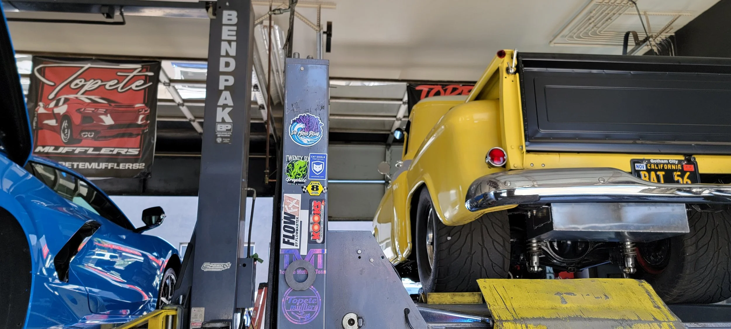 Close-up of a yellow vintage pickup truck lifted on a car shop lift, with various automotive stickers on the lift and a blue sports car partially visible on the left.