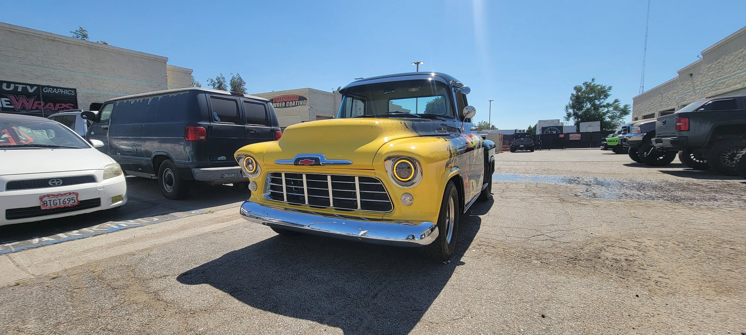 A yellow vintage car with a black roof, parked among other modern vehicles in a parking lot on a sunny day.