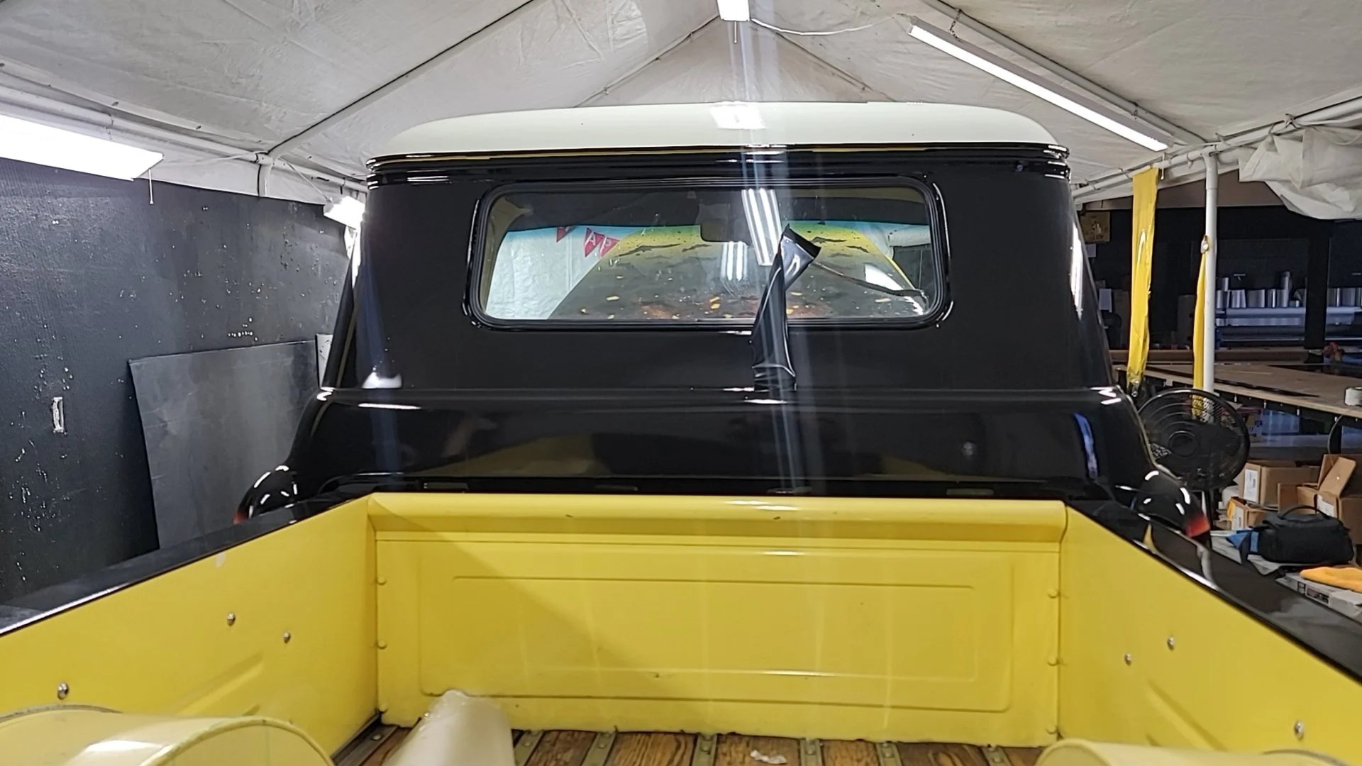 The image shows the front view of a vintage vehicle, possibly a small truck or utility truck, inside a garage or workshop with a black upper body and a white roof, behind a yellow barrier or platform.