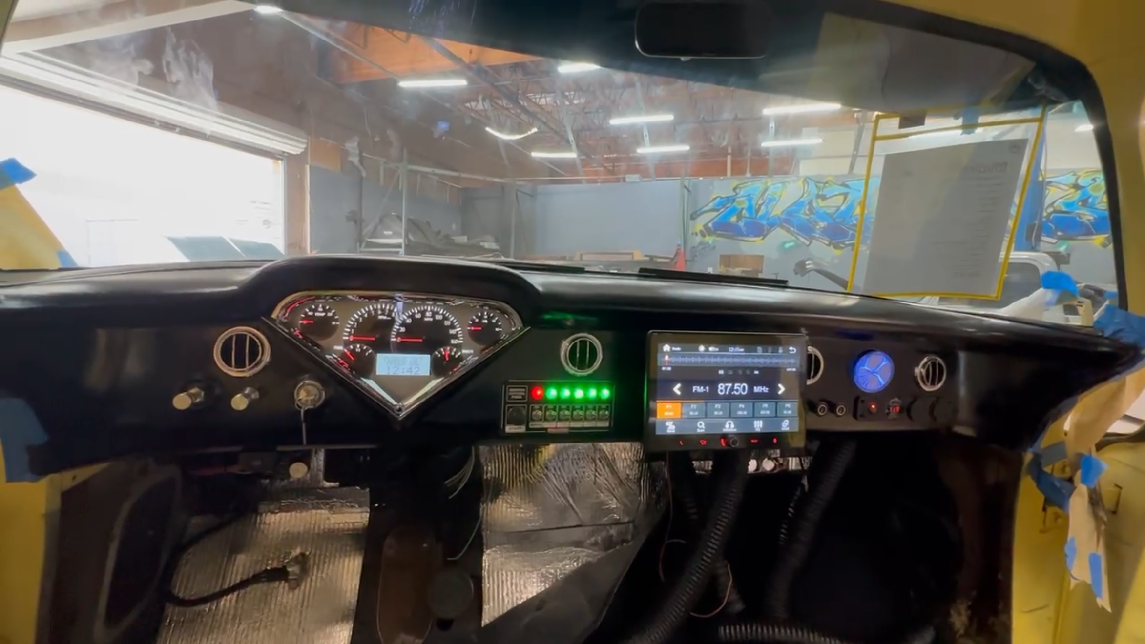View of vehicle dashboard with gauges, radio, and control panel in a workshop or garage with bright lighting and graffitied wall in background.