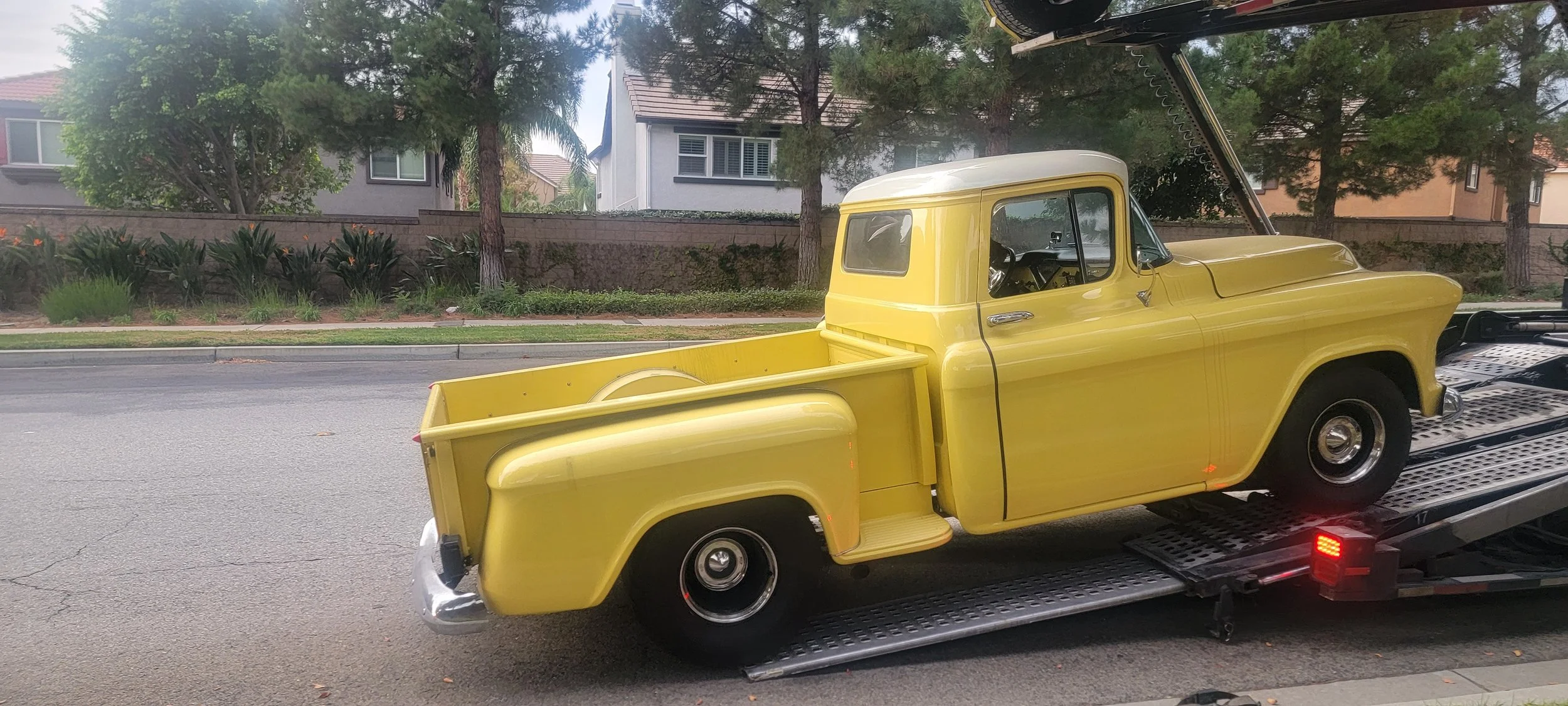 Yellow vintage pickup truck being transported on a flatbed tow truck in a residential neighborhood with trees and houses in the background.