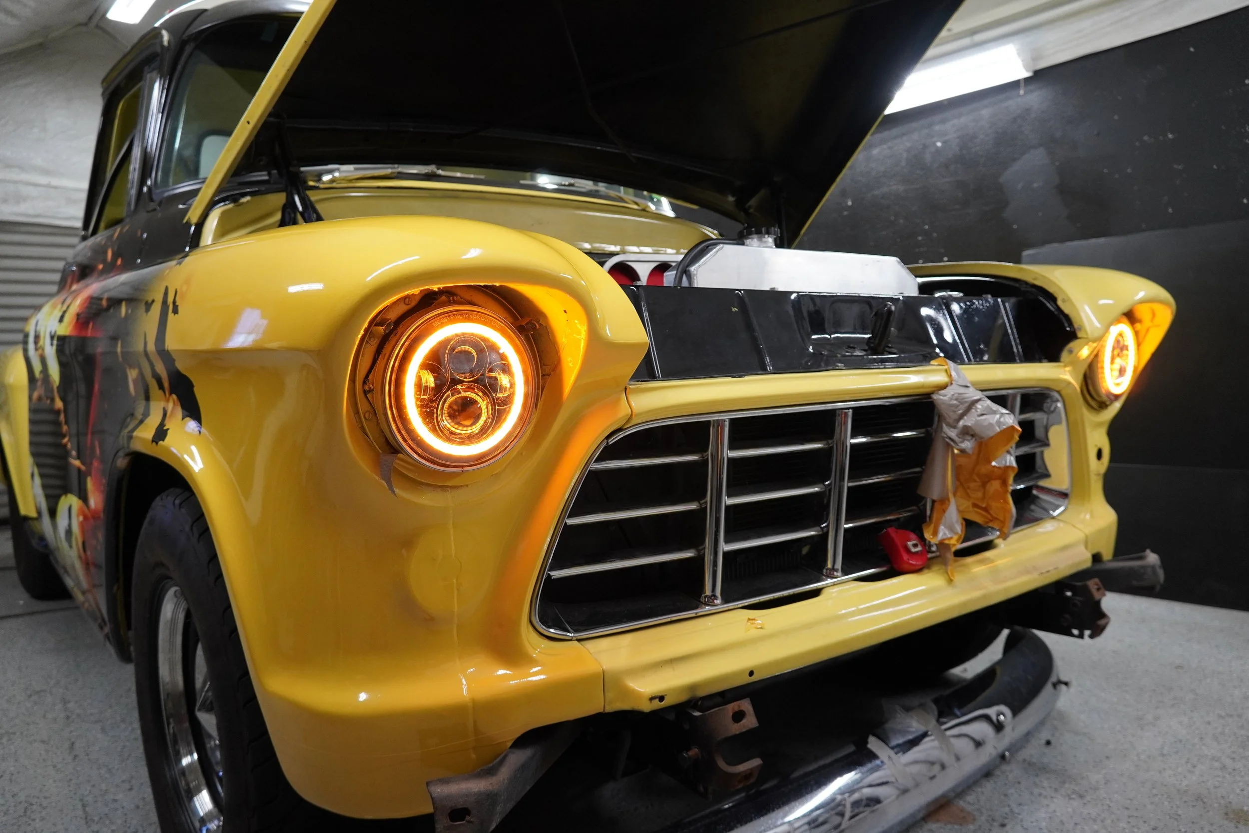 A yellow classic car with its hood open in a garage, showing the engine compartment and headlight, in the process of restoration or repair.