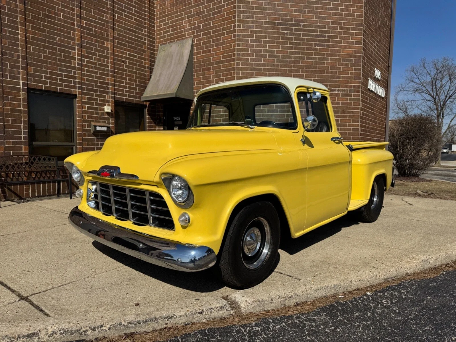 A vintage yellow pickup truck parked outside a brick building on a sunny day.