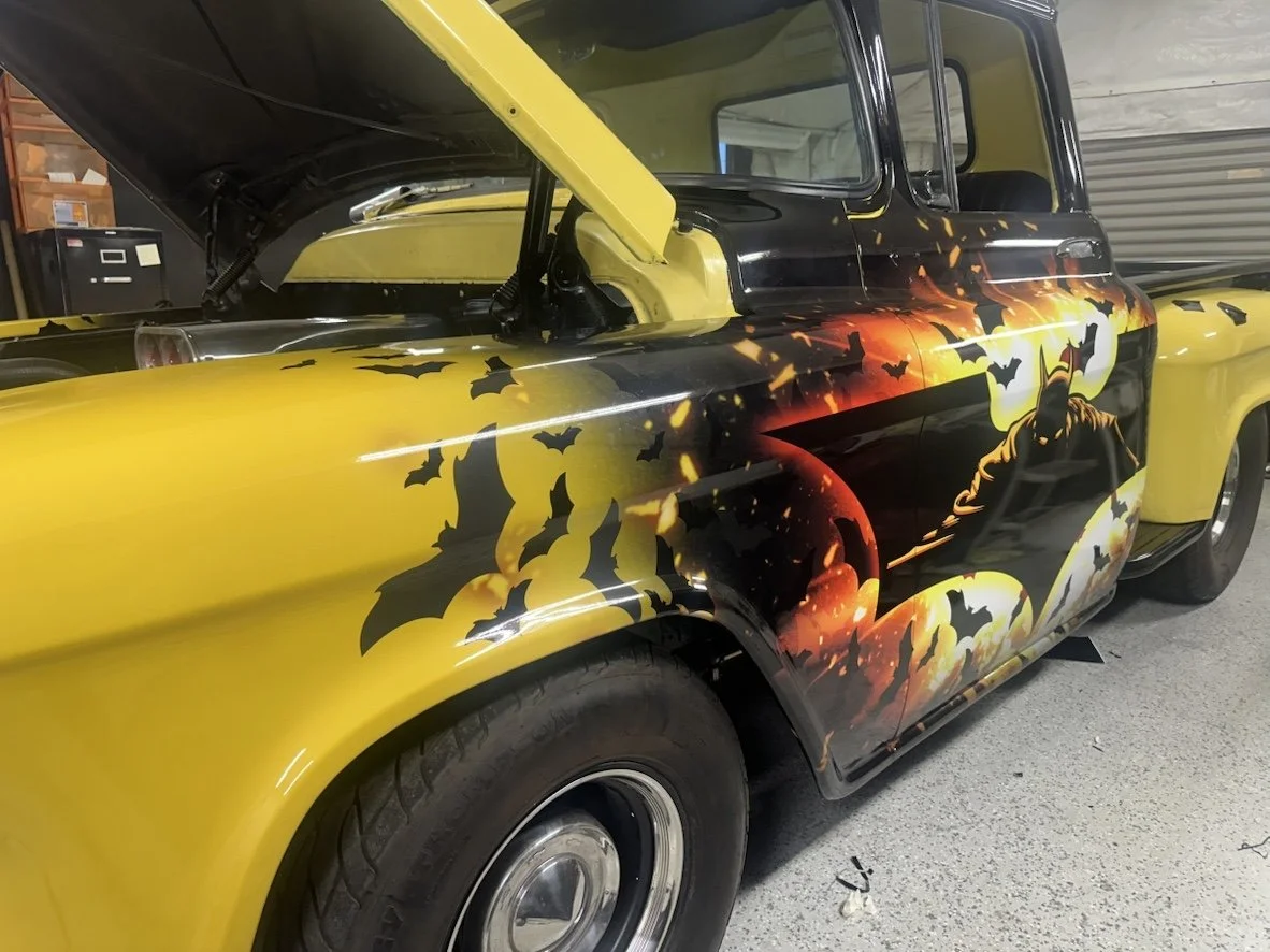 Yellow car with custom batman-themed artwork featuring bats, a full moon, and Batman on the side, parked in a garage with its hood open.