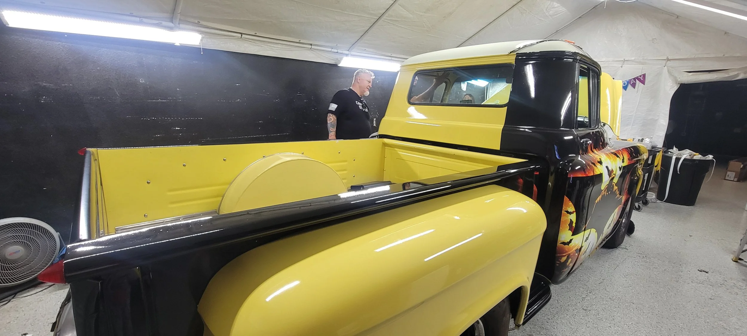 A yellow and black vintage pickup truck with flame artwork parked inside a large tent, with a man with tattoos standing nearby.