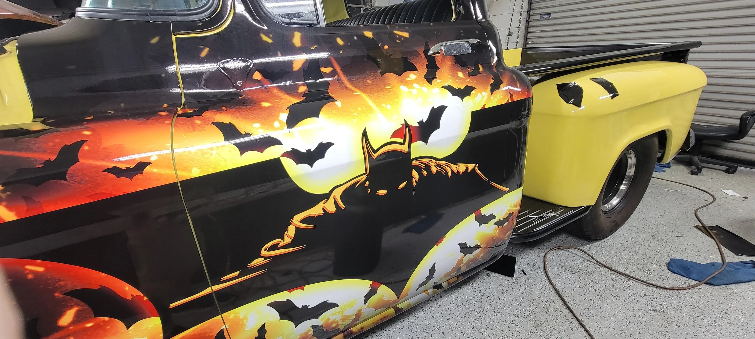 A classic pickup truck with a Halloween-themed custom paint job featuring bats, a large full moon, and a figure in a witch's hat, parked in a garage. The truck's body is primarily black with bright yellow accents.