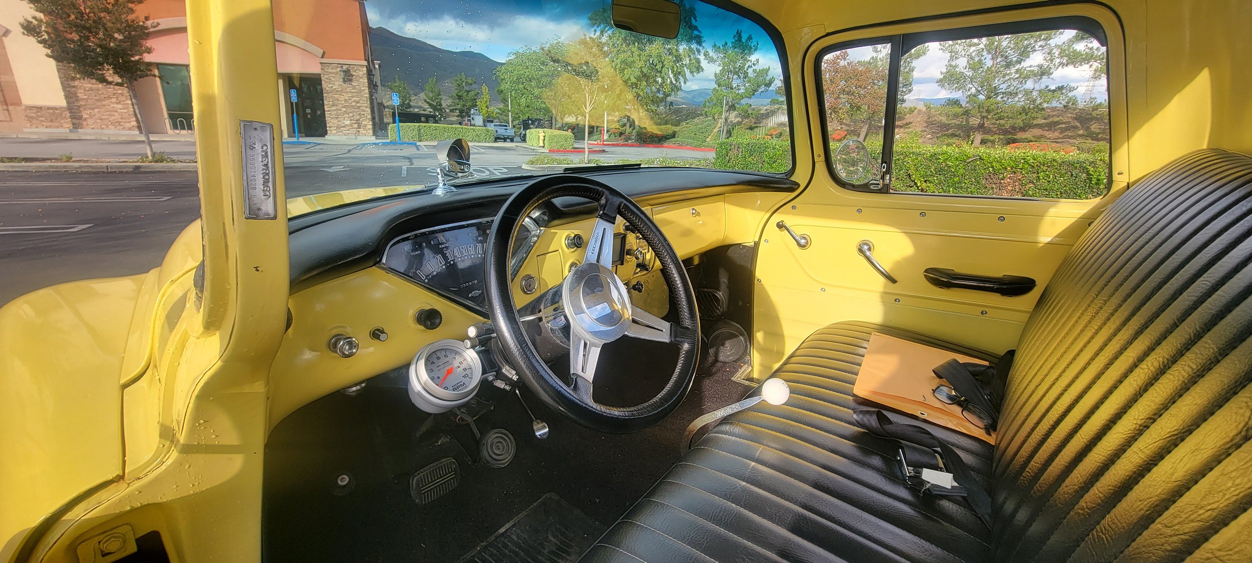 Interior view of a vintage yellow truck showing the steering wheel, dashboard with speedometer and other gauges, gear shift, and a bench seat with a notebook, spoon, and sunglasses on it. Through the windshield, a parking lot with trees, bushes, hill