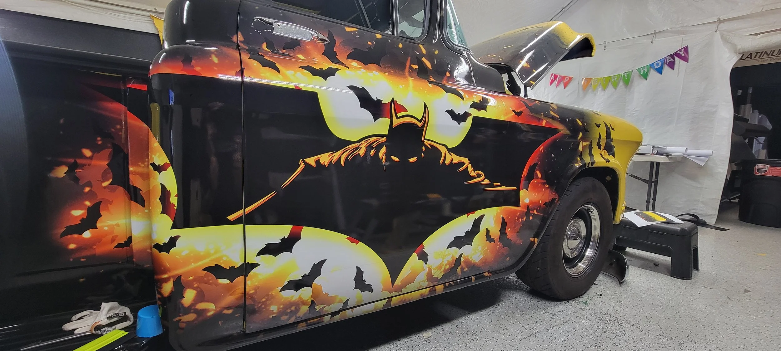 A black vintage car with Batman-themed custom artwork, featuring Batman's silhouette and bats against a fiery yellow and orange background.