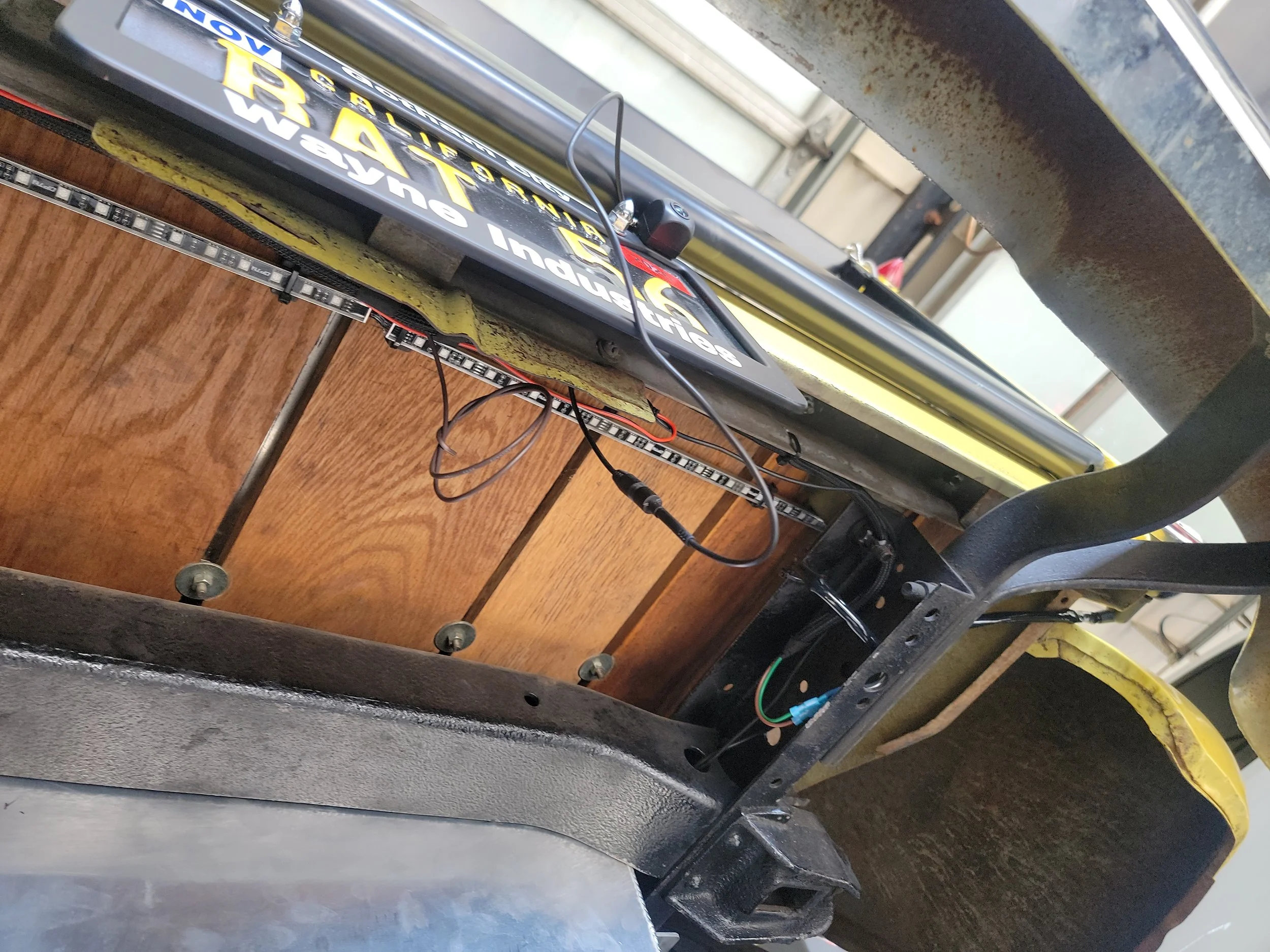 Close-up of a black bicycle frame with stickers and wires, placed on a wooden surface with a metal and yellow frame nearby.