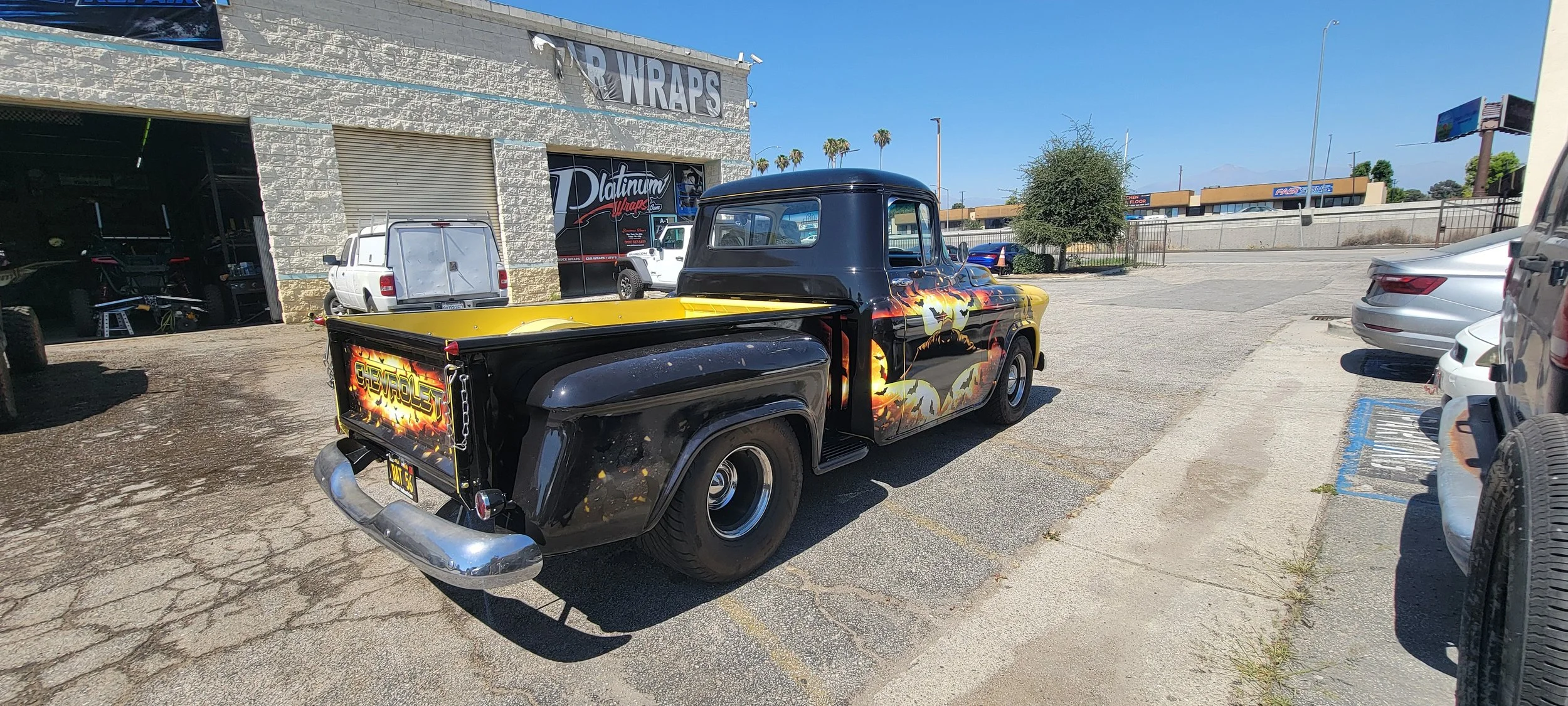 A vintage black pickup truck with flame and skull artwork parked in a commercial parking lot.