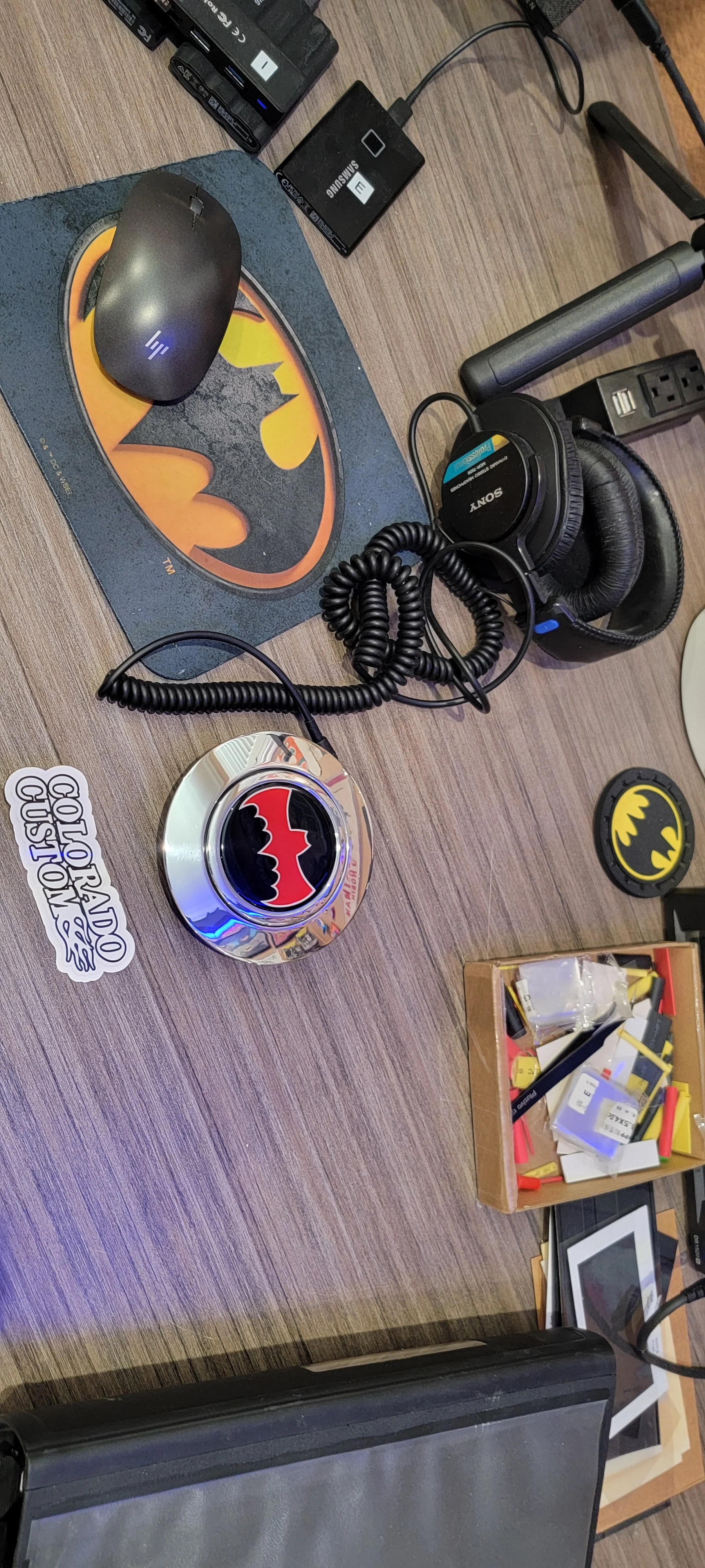 Desk with Batman-themed items, including a Batman sticker, Batman circular badge, Batman patch, and Batman mouse pad. There are also electronic accessories like headphones, batteries, and small tech parts scattered on the surface.
