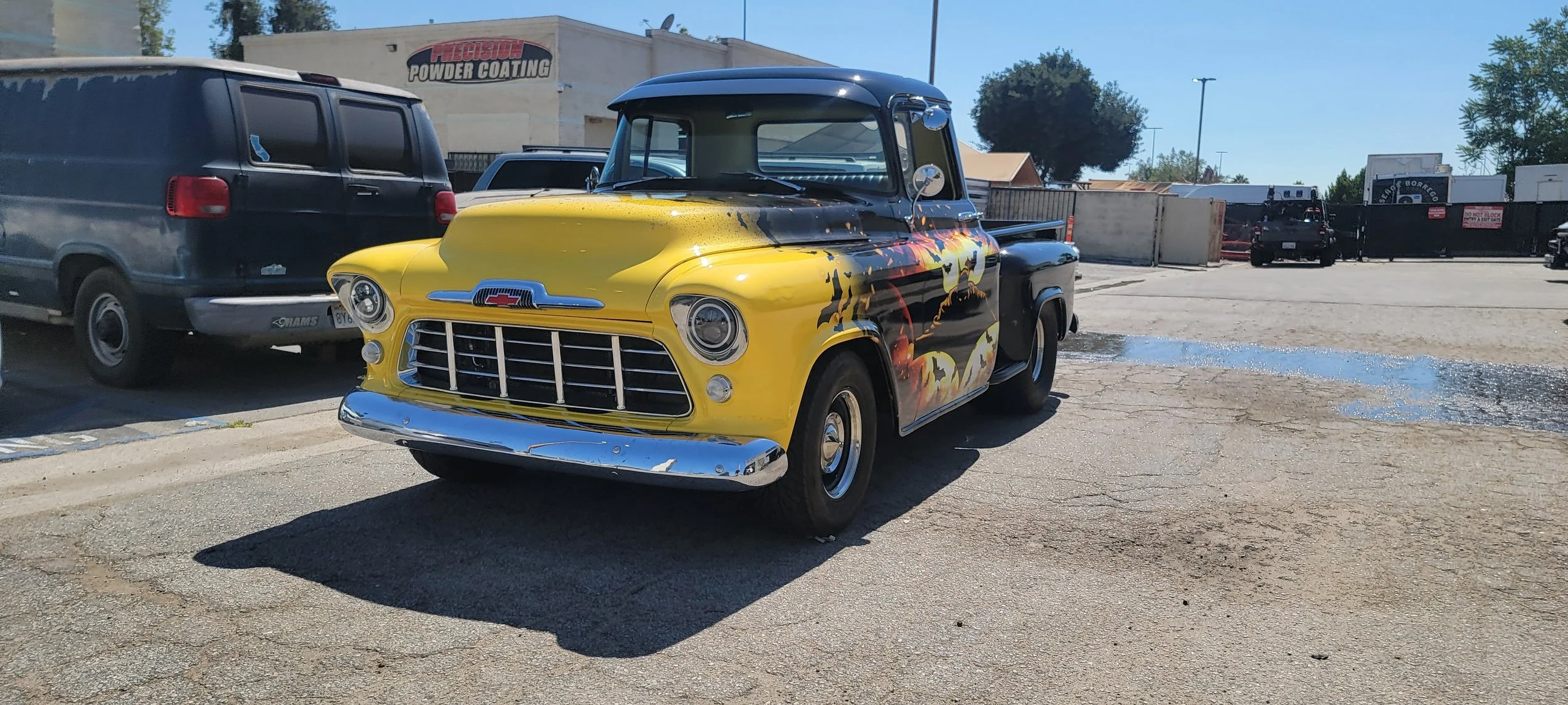 Vintage pickup truck with yellow and black paint featuring a flaming pumpkin design, parked in a parking lot next to other vehicles.