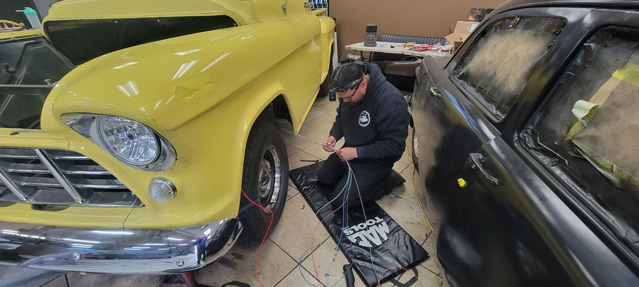 A man working on a vintage yellow car in a garage, wearing a headlamp, with multiple car parts and tools around him.