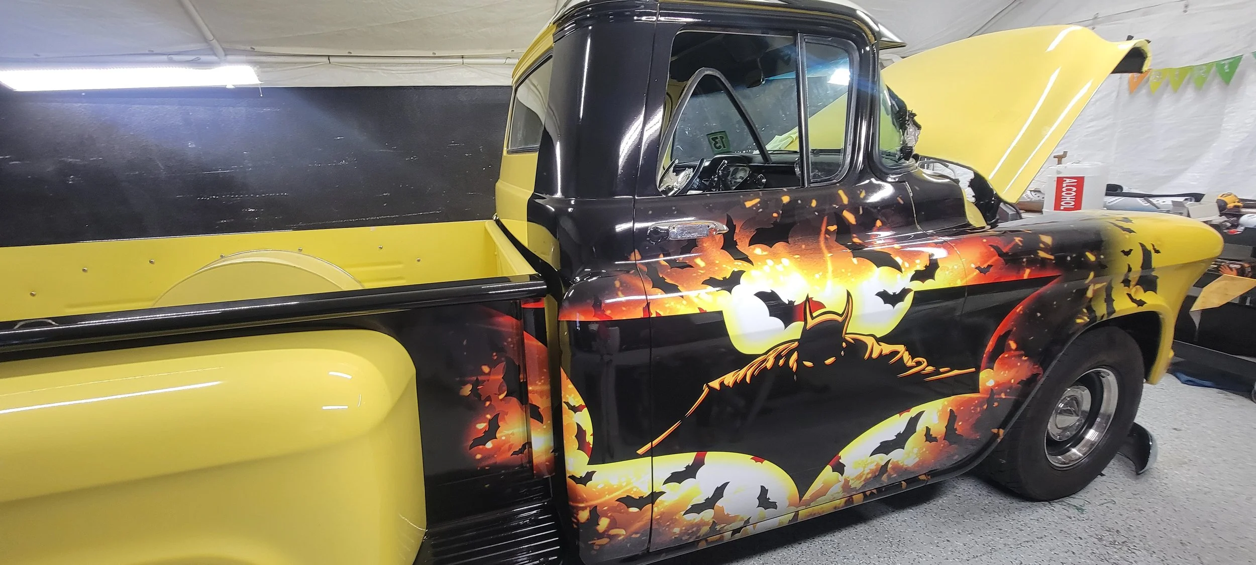 A vintage pickup truck with a Batman-themed airbrushed paint job featuring bats, a full moon, and Batman's silhouette in front of an orange and black background.