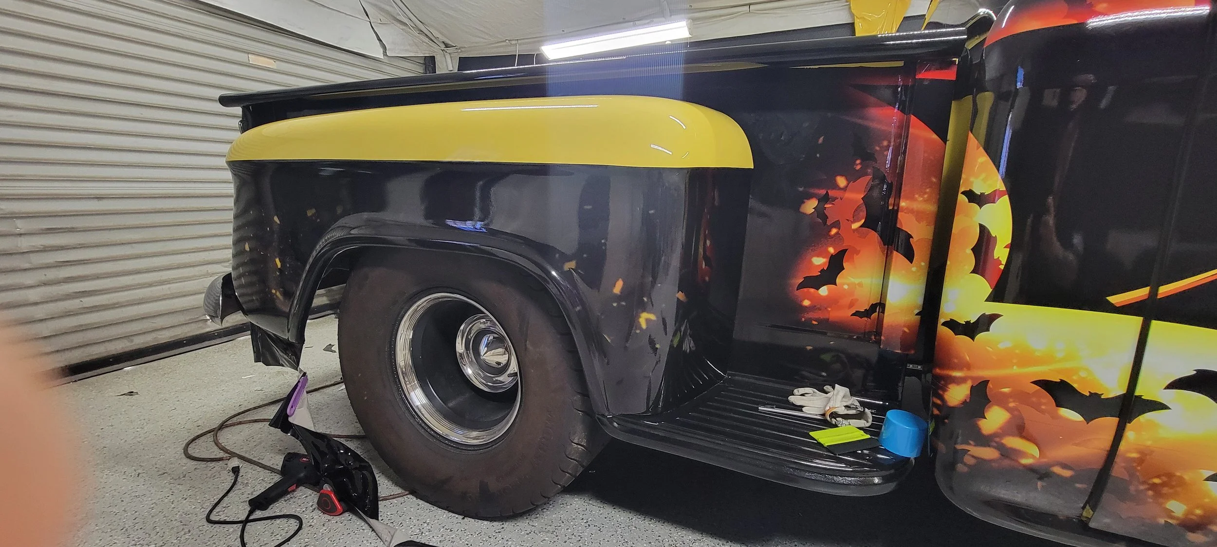 A vintage black truck with a Halloween-themed wrap featuring bats and a sunset background, parked inside a garage.