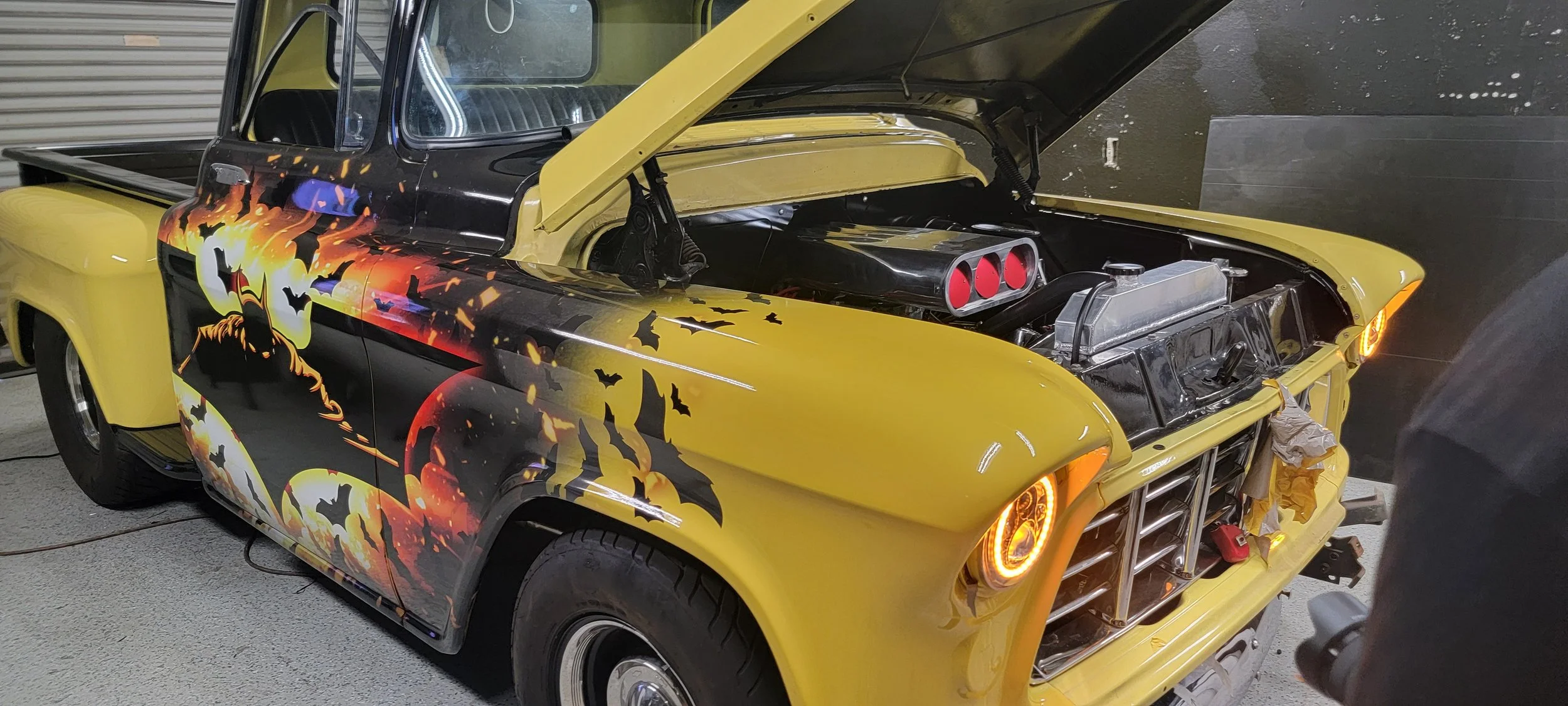 A yellow pickup truck with a painted Halloween-themed design featuring pumpkins, bats, and a haunted house. The hood is open, showing the engine, with parts being worked on in a garage.