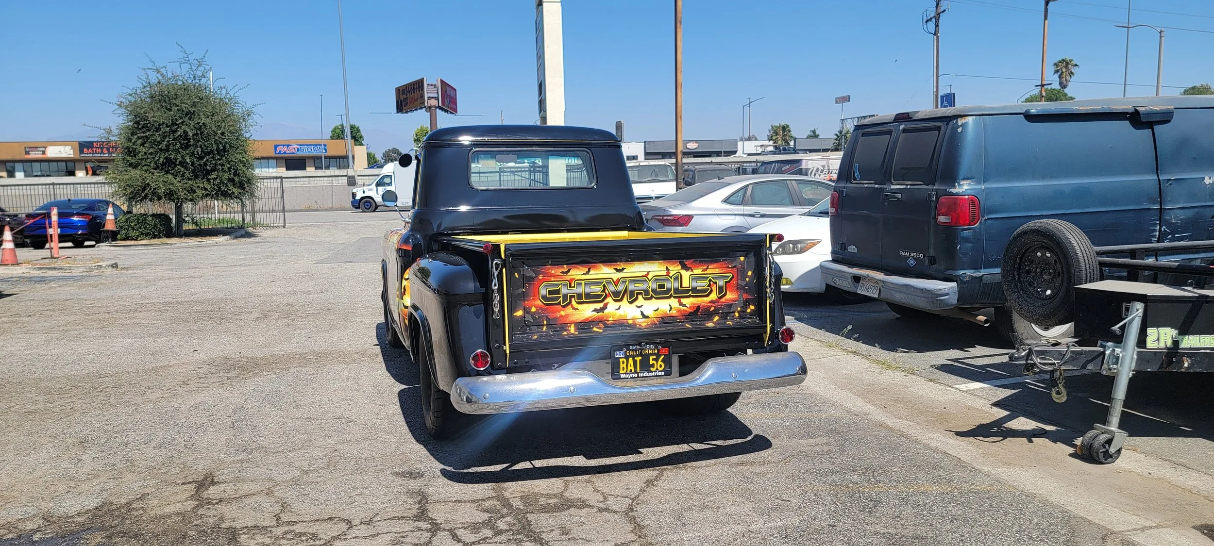 A vintage Chevrolet pickup truck with custom flame paint and a license plate that reads BAT 56, parked in a lot with other vehicles. The truck has a black cab and a dramatic, fiery graphic on the tailgate.