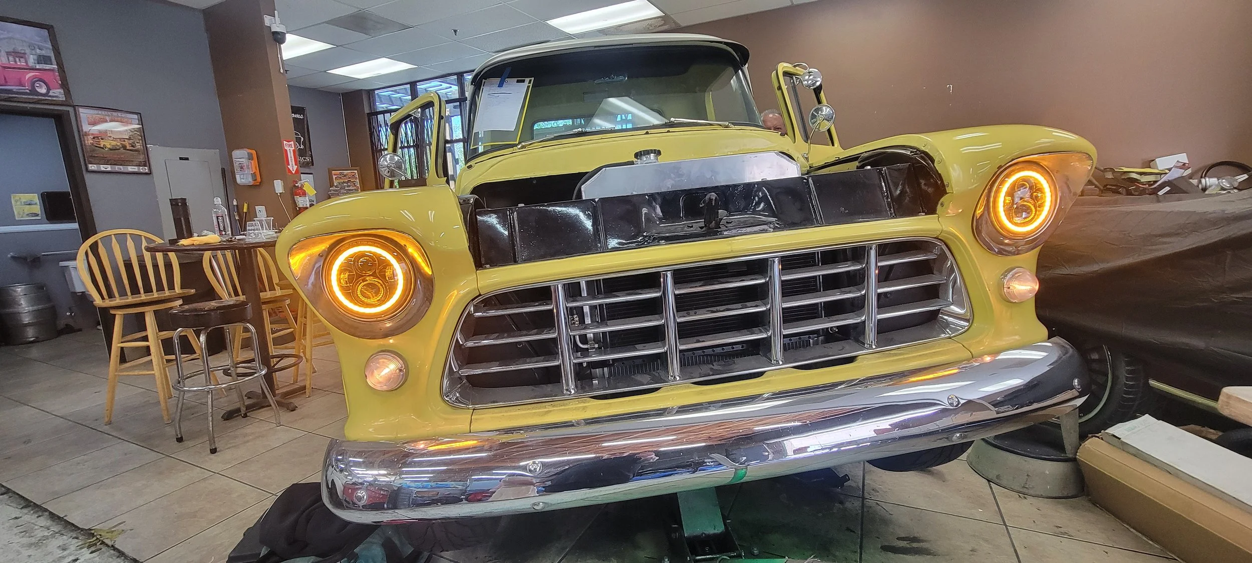 A partially assembled yellow vintage car inside a workshop, with its front grille, headlights, and hood area visible, surrounded by various tools and furniture.