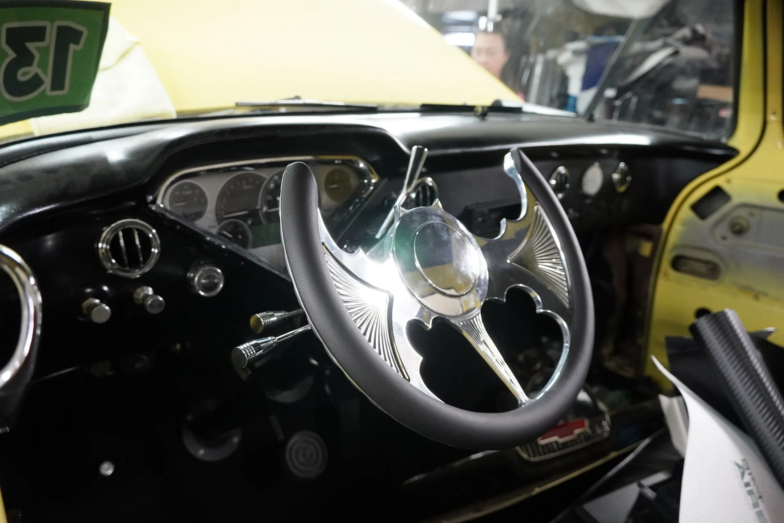 Close-up of a vintage car dashboard, steering wheel with a metallic decorative center, and various gauges and controls, with a yellow exterior visible through the windshield.
