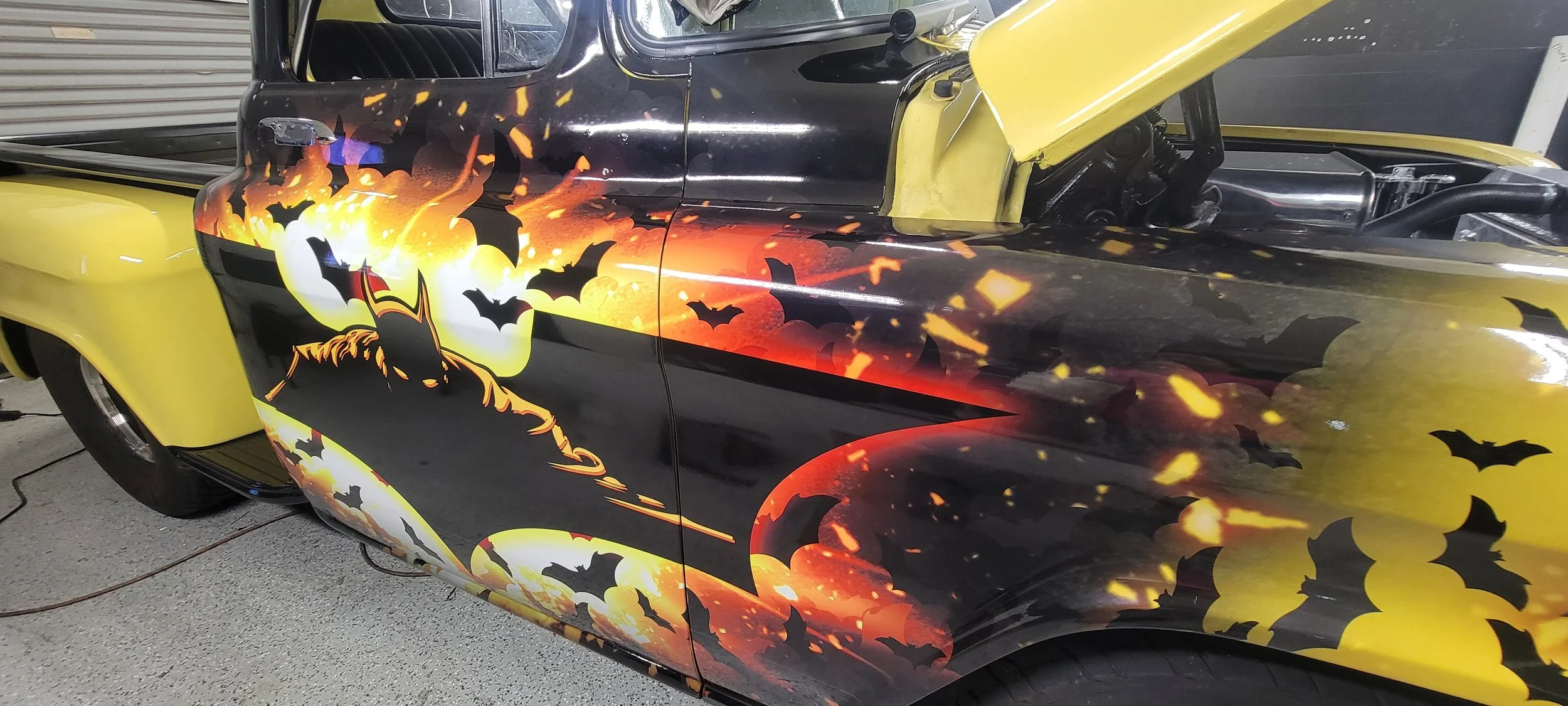 A vintage car with a Halloween-themed paint job featuring a large moon, bats, and fiery colors, with yellow accents on the fenders.