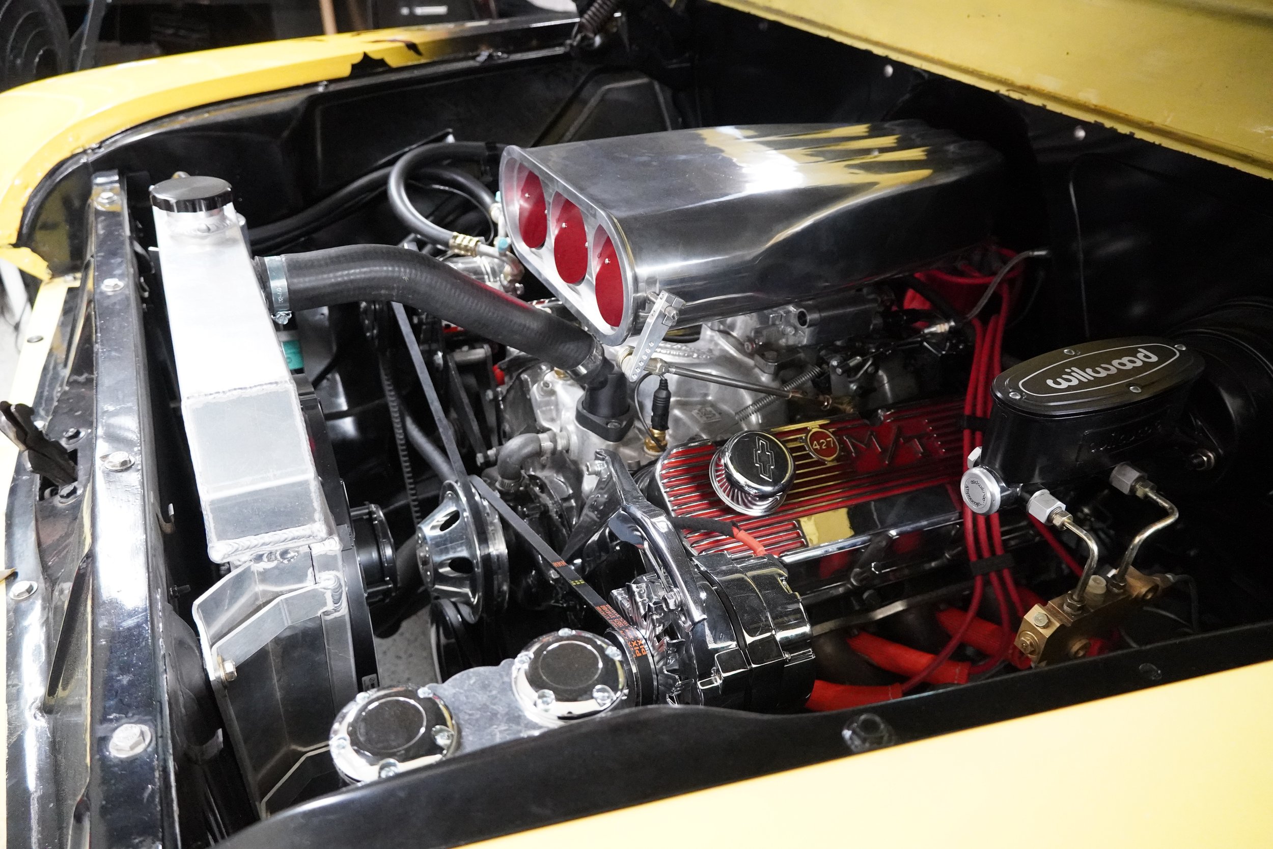 Close-up of a race car engine with polished metal parts, including a large intake manifold, with red spark plug wires and various hoses and fittings visible.