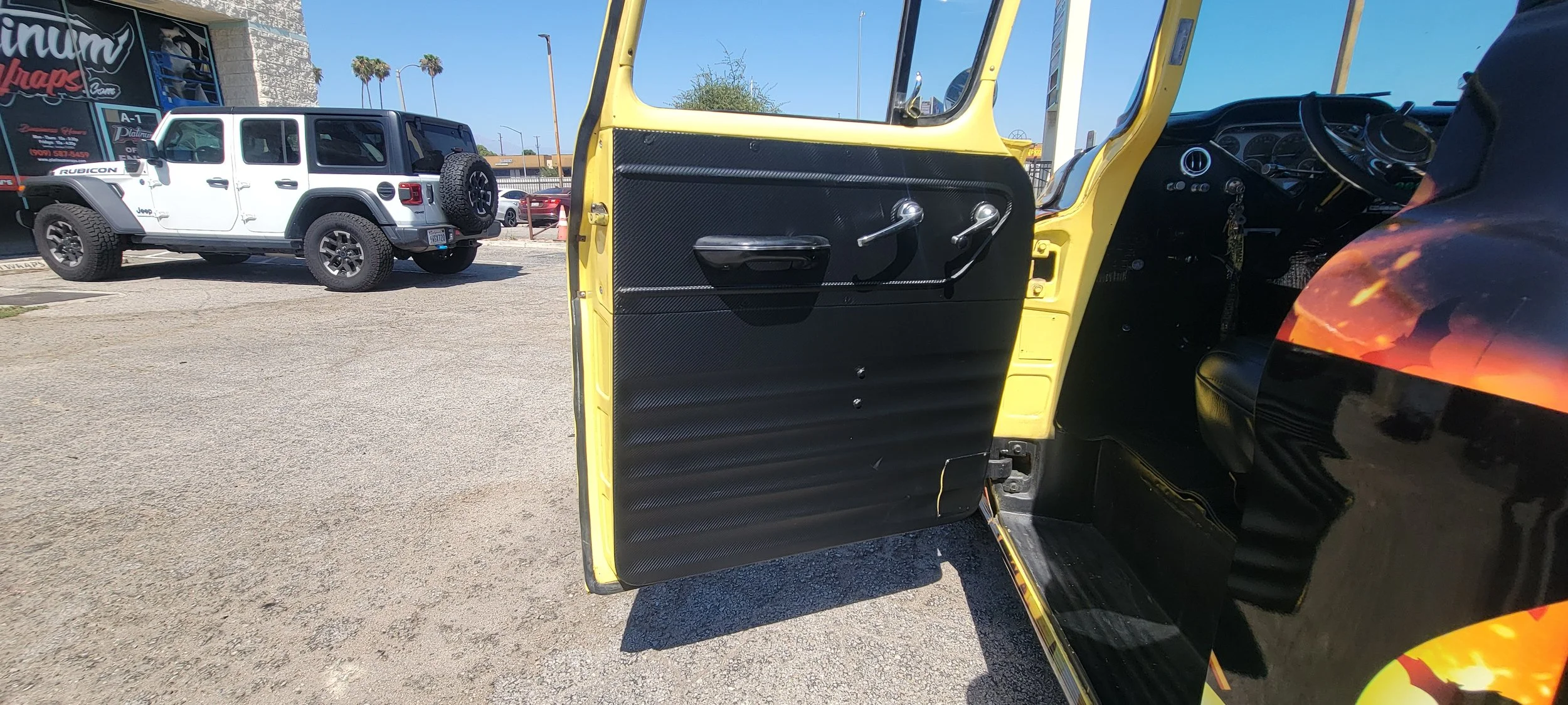 A black vehicle with a carbon fiber inside door panel, a yellow exterior, and a black steering wheel. Outside, a white Jeep Rubicon with a spare tire mounted on the back is parked across a paved lot, with part of a black and red sign and some palm tr