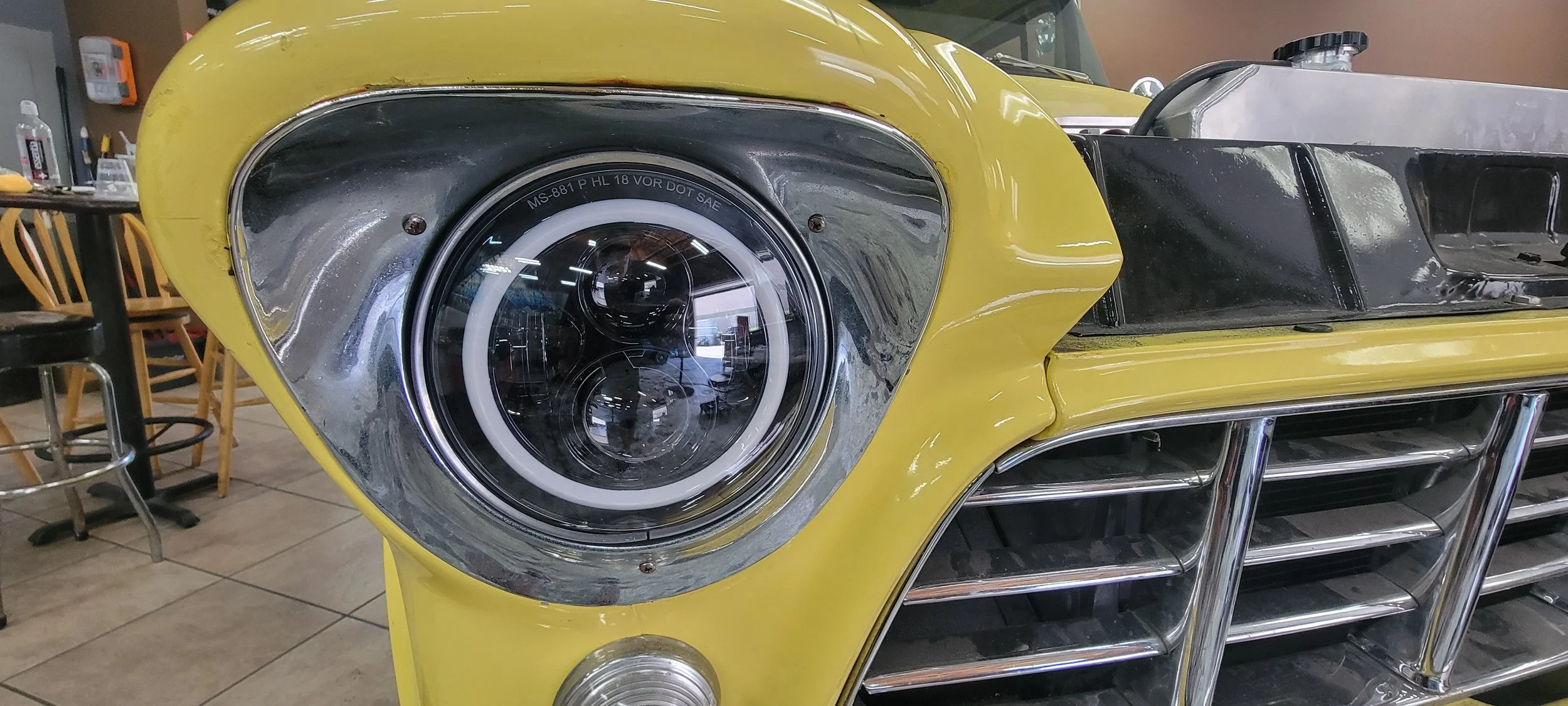 Close-up of the front of a yellow vintage car with a chrome headlight and grille, inside a showroom or workshop.