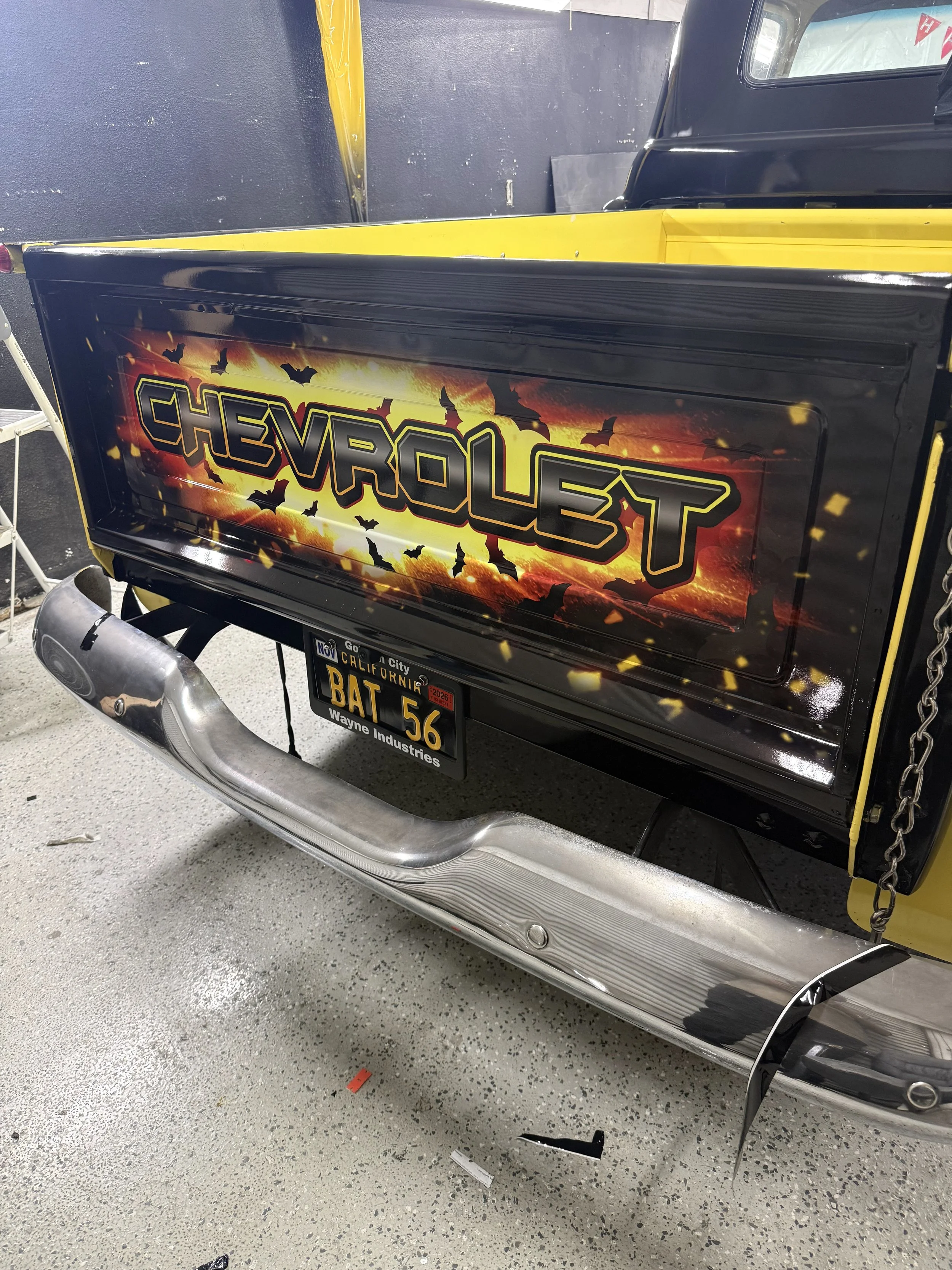 The rear of a black and yellow Chevrolet vehicle, possibly a vintage or custom car, with a graffiti-style design on the side that says 'CHEVROLET' and features bat silhouettes. The vehicle has a California license plate reading 'BAT 56' and is parked