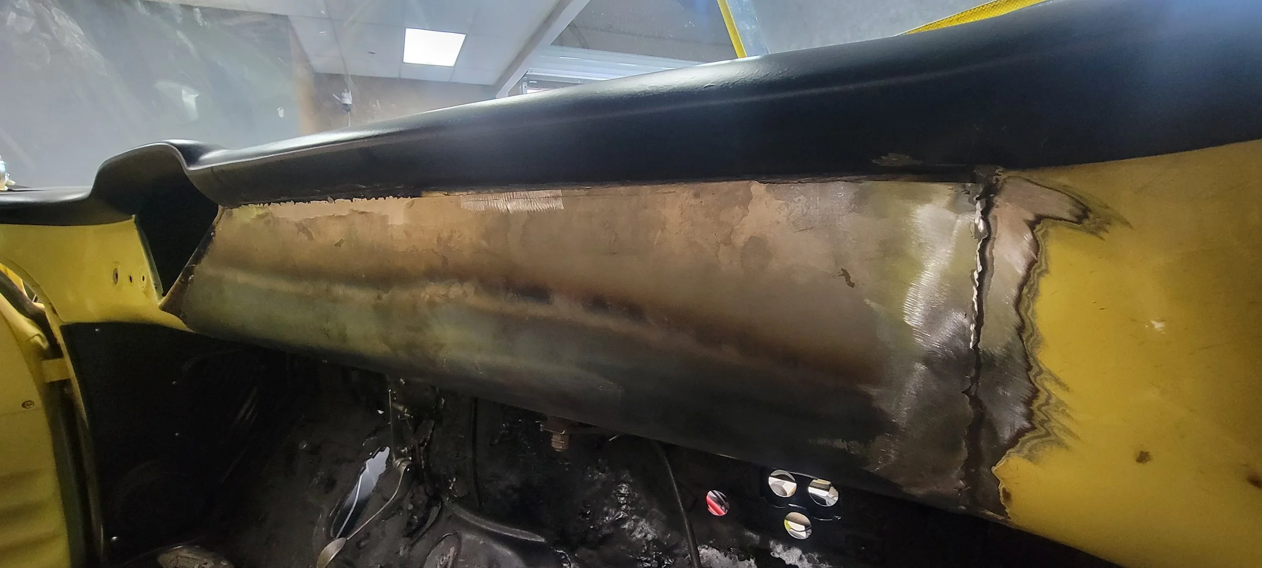 Interior view of a vehicle with a large, burned oval-shaped hole in the metal floor and side panels, exposing internal structures and wiring.
