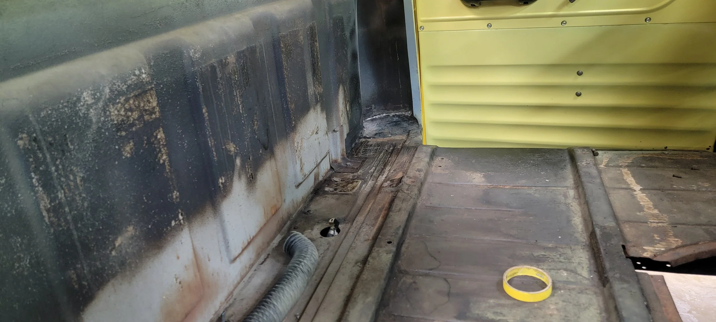 An industrial workspace with a dirty metallic surface, a flexible hose, a yellow wall with some bolts, and a yellow roll of tape on a wooden surface.