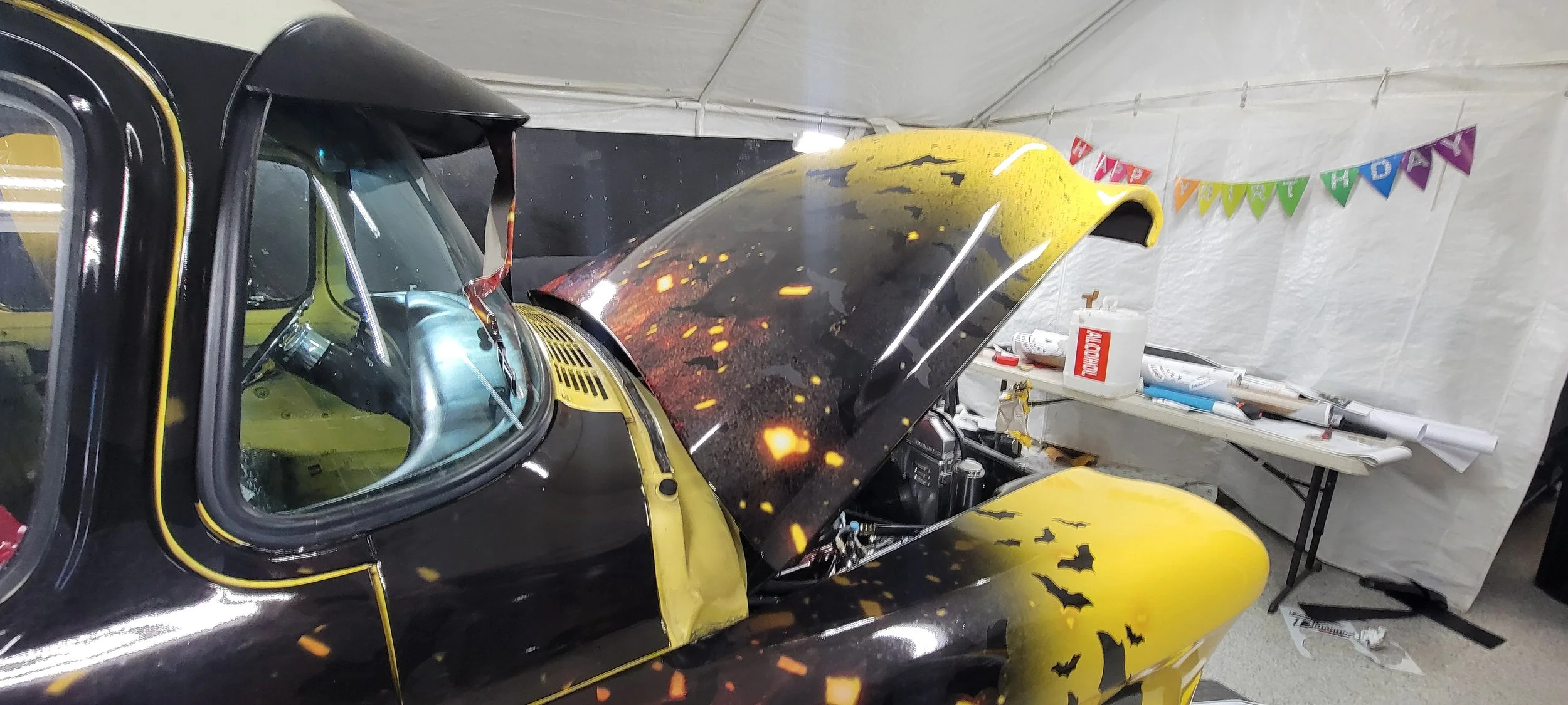 A yellow and black race car with a Batman logo on the side in a workshop with a banner reading 'Happy Birthday'.