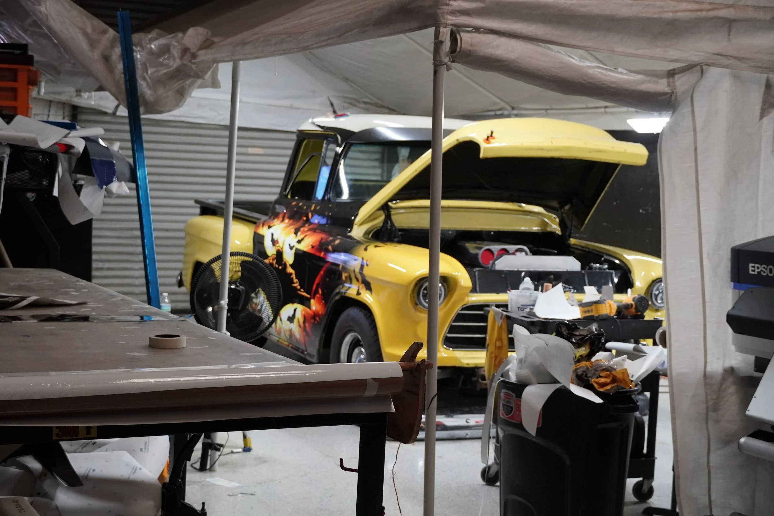 Garage with a yellow car that has a black hood and flaming decals, with the hood open, surrounded by tools and equipment.