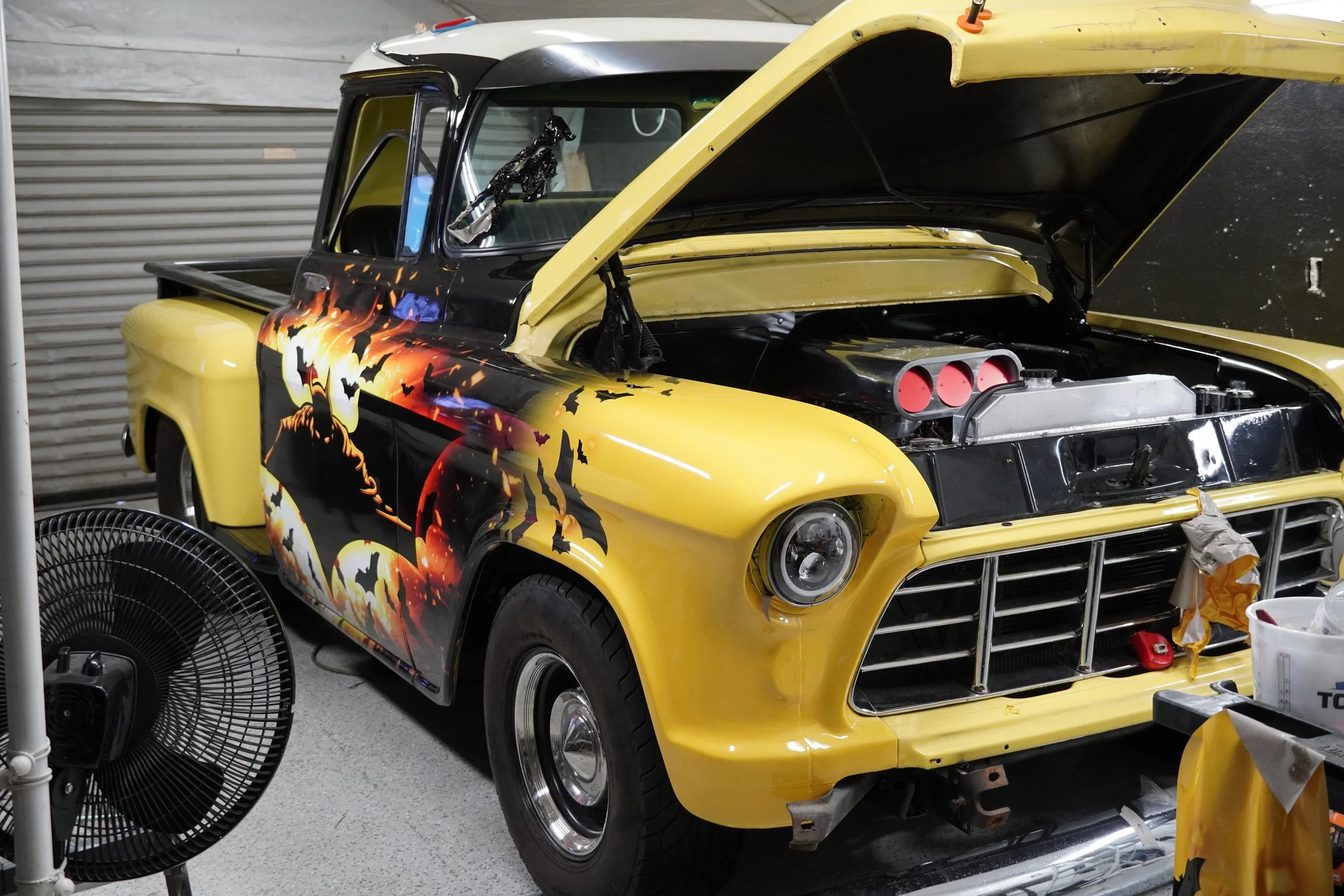 A vintage pickup truck with a Halloween-themed design featuring bats and a pumpkin on its side, parked indoors with its hood open.