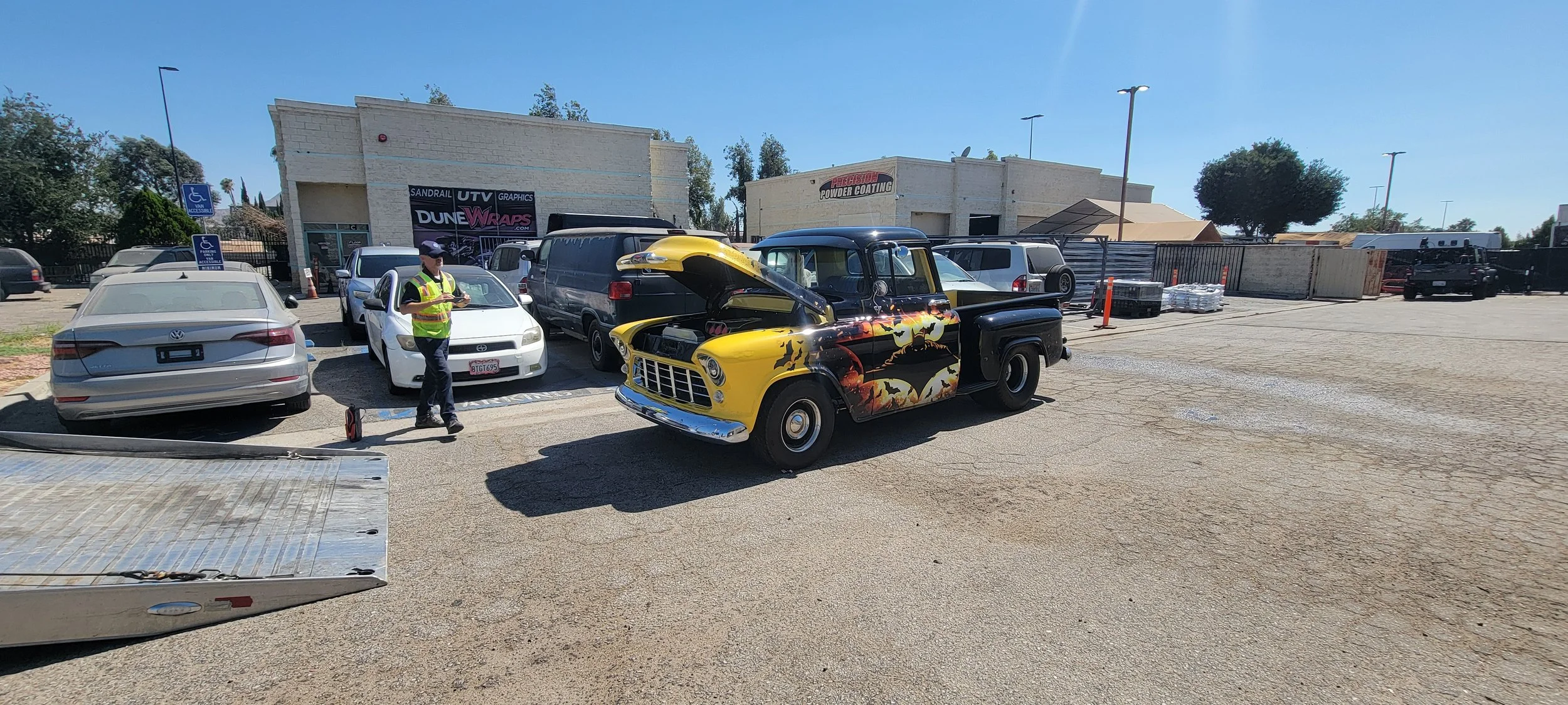 A parking lot with several parked cars and a vintage yellow and black pickup truck with a dragon design on its side. A man in a yellow safety vest and cap is standing next to a white car, looking at a device. In the background, there are buildings wi