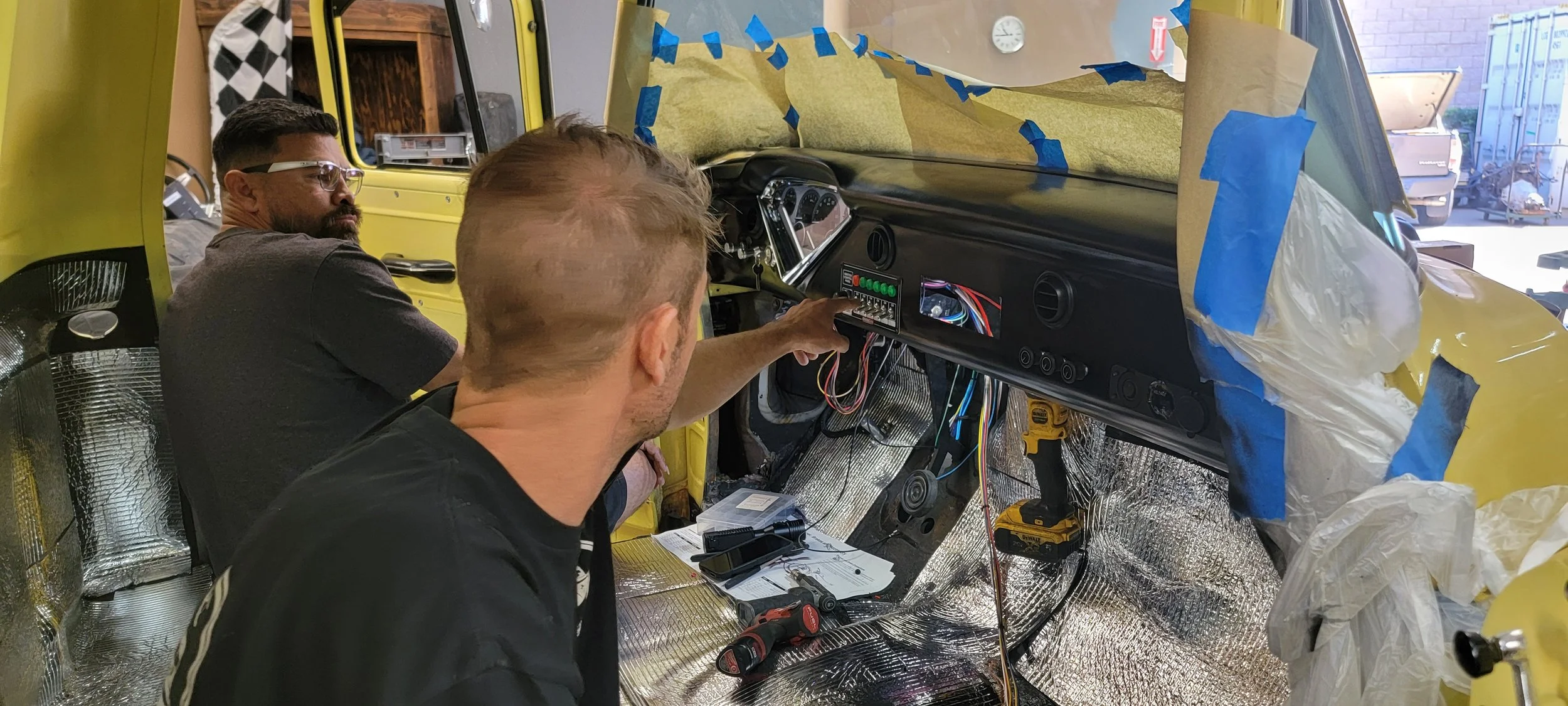 Two men working on installing or repairing the dashboard of a yellow car in a workshop, with blue painter's tape and insulation material inside the car and tools on the floor.